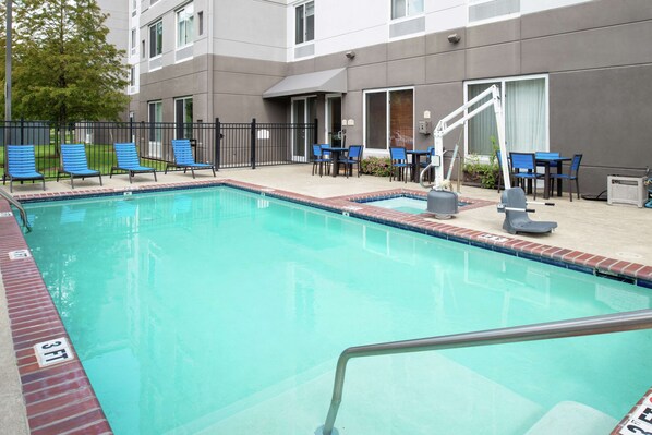 A swimming pool with blue chairs and tables, surrounded by a building with a grey facade and a black fence.
