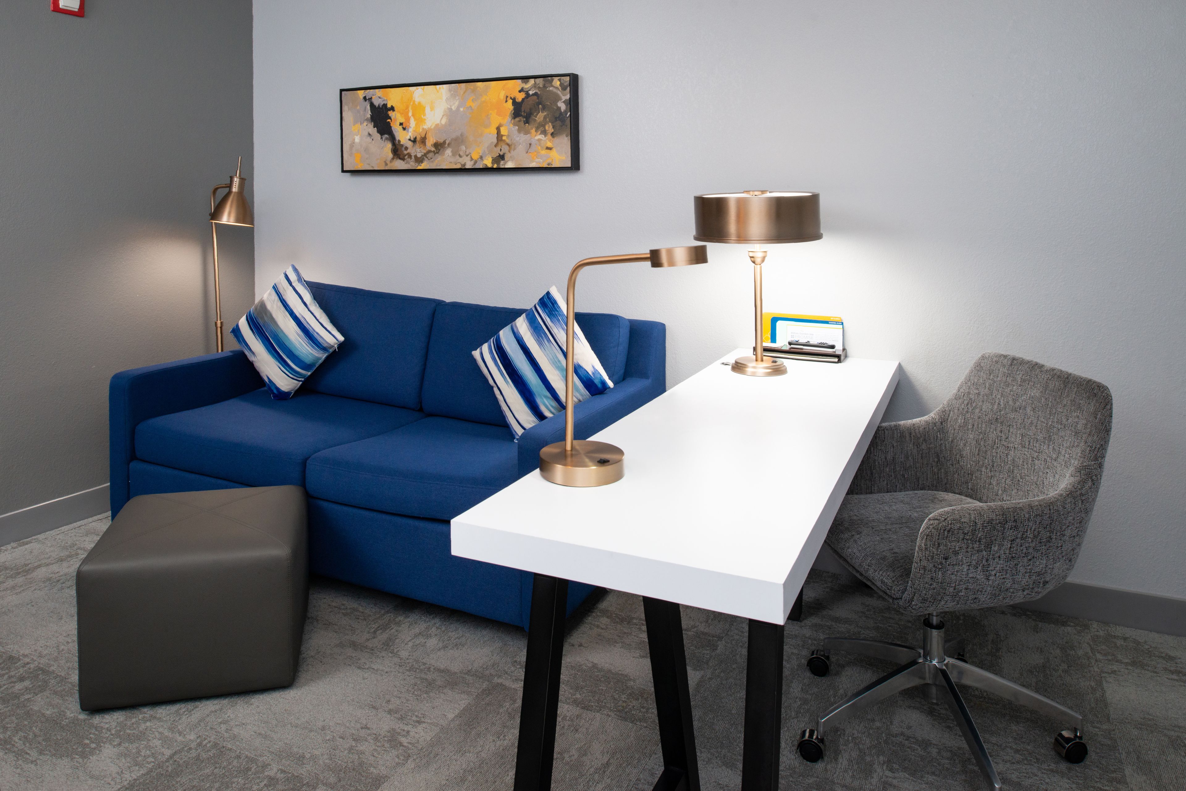 Ein modernes Zimmer mit einem blauen Sofa, einem weißen Schreibtisch, einem grauen Bürostuhl und einem kleinen grauen Hocker.