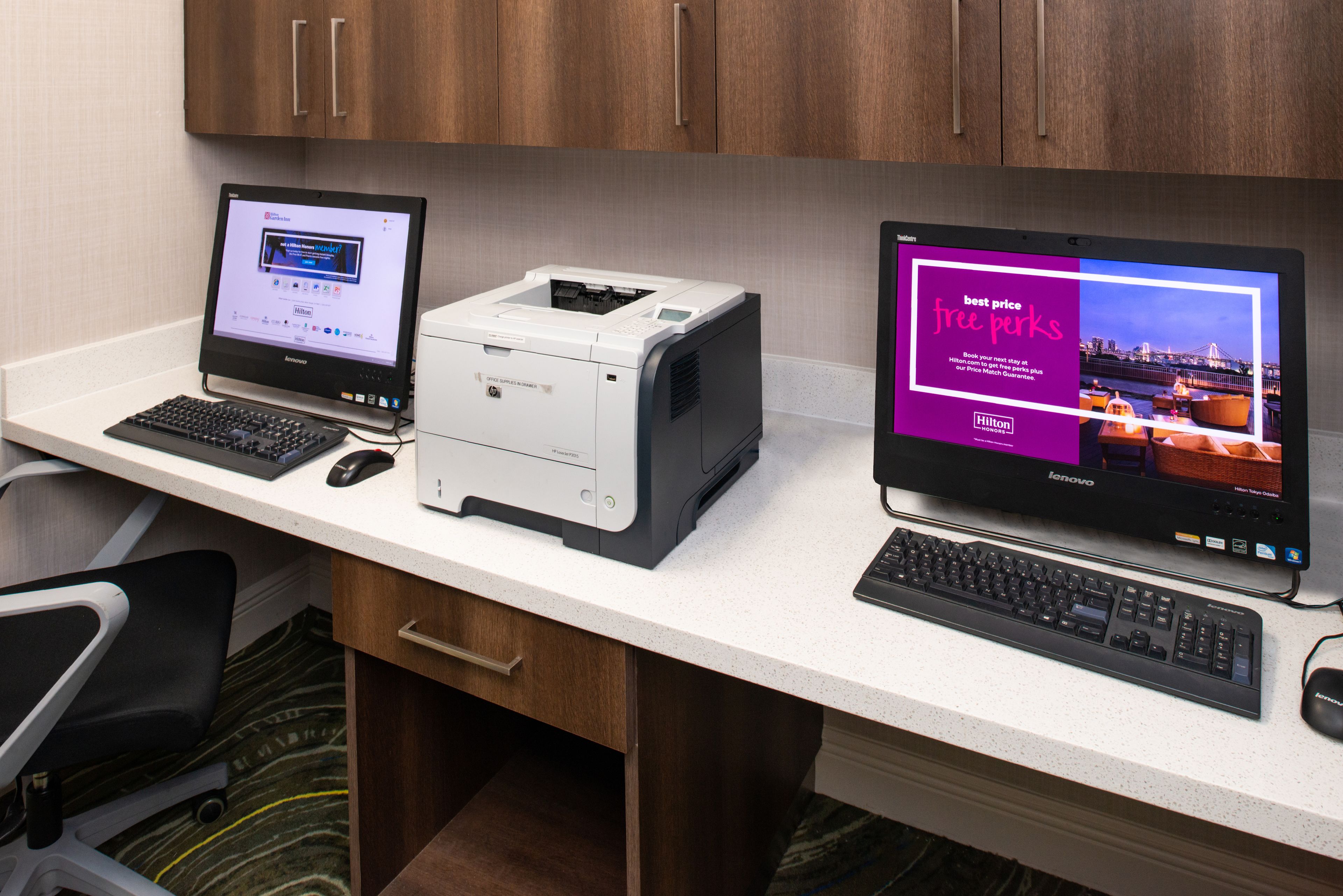 A hotel room desk with two monitors, a printer, and a keyboard.