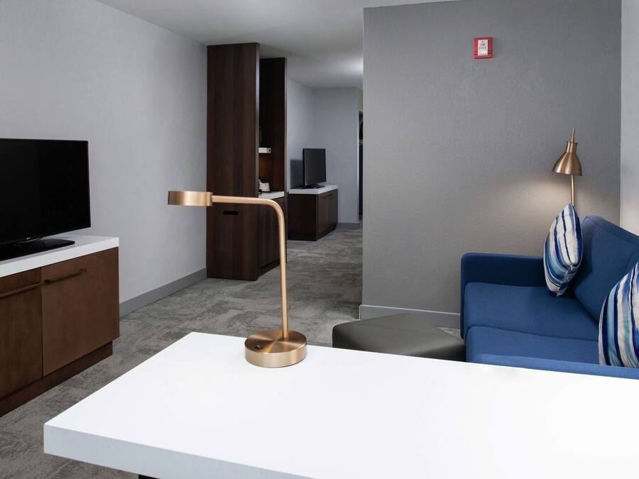 A modern living room with a flat-screen TV on a white cabinet, a blue sofa with striped cushions, a gold floor lamp, and a grey wall with a red fire alarm.