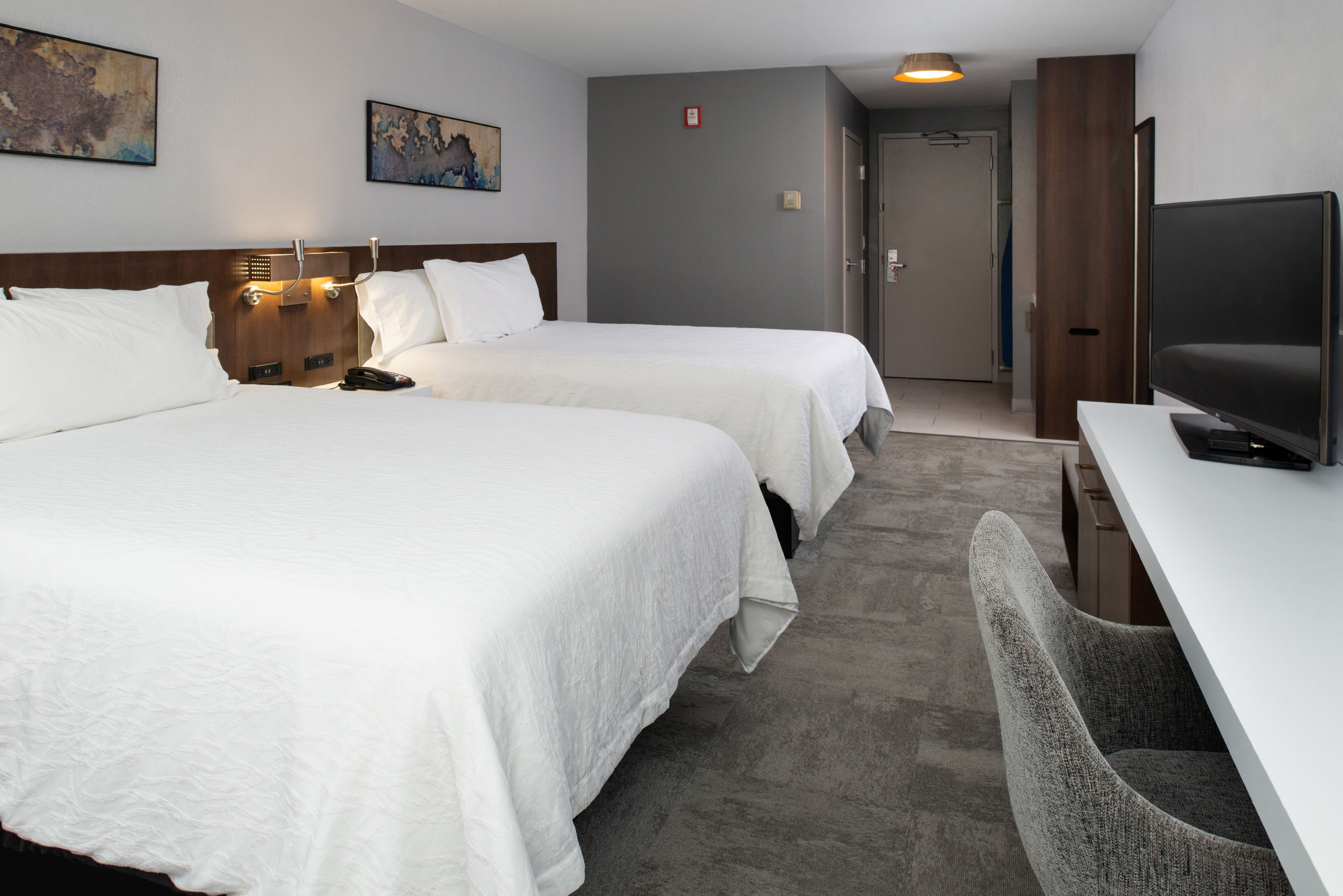 A hotel room with two beds, a desk, a television, and artwork on the walls.