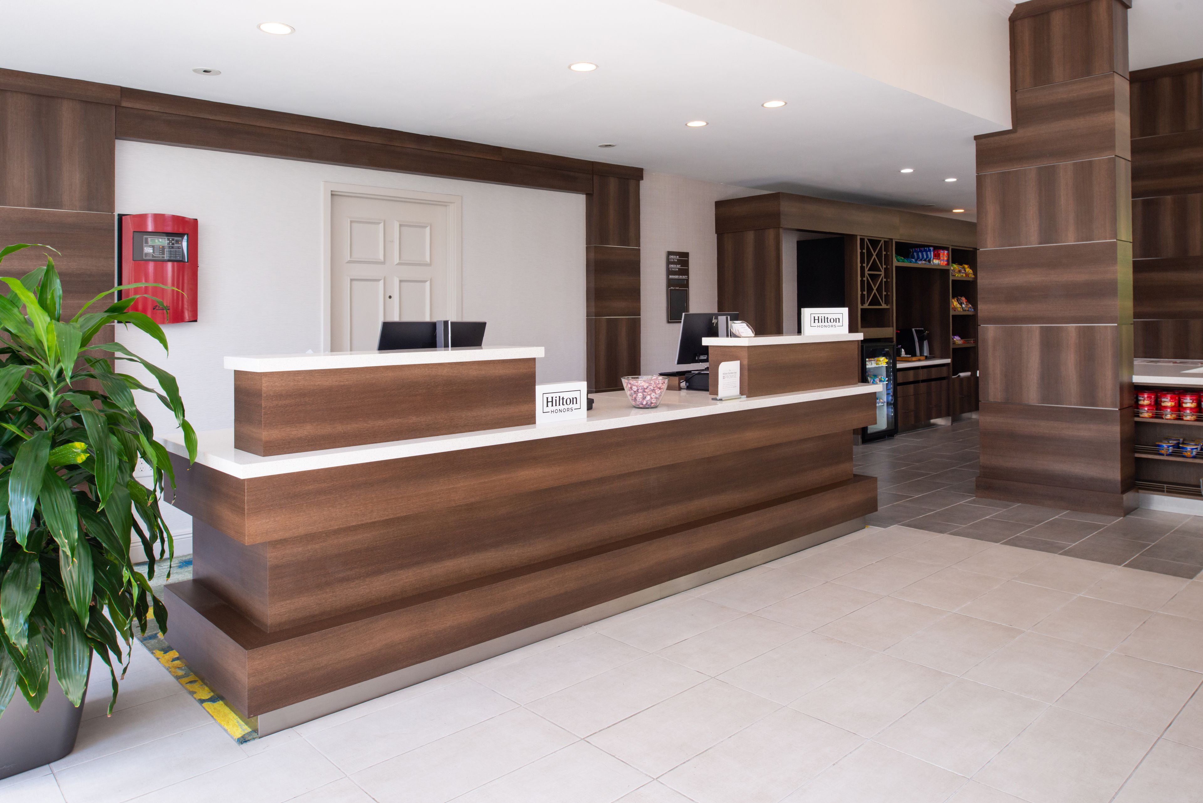A modern hotel reception desk with a wooden finish, a white door, and a potted plant to the left.