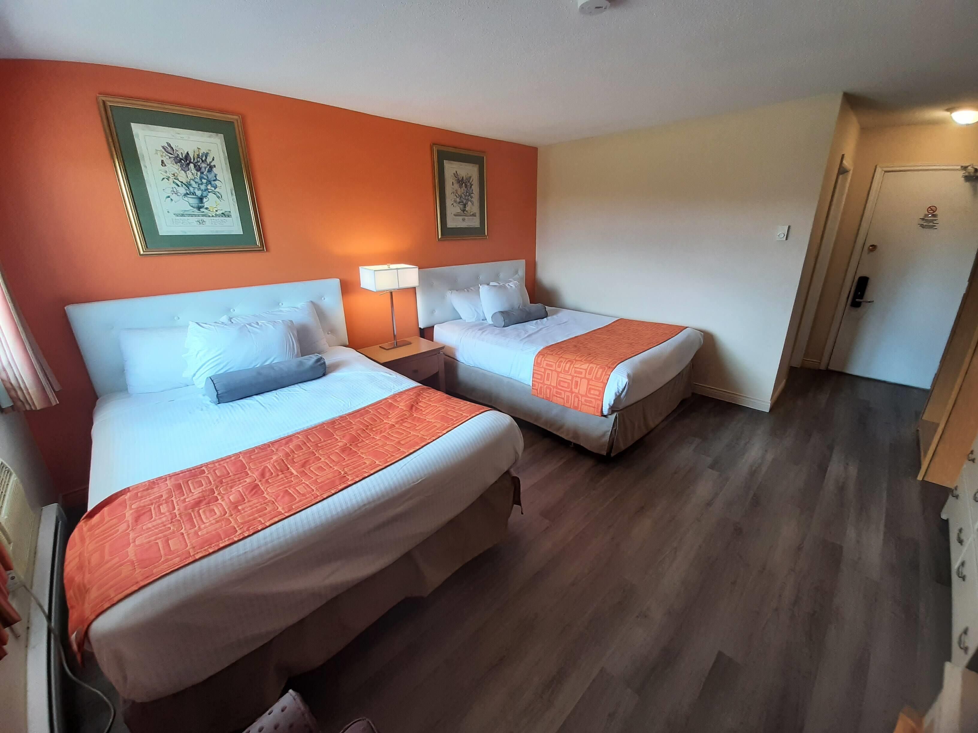 A hotel room with two beds, each with a white headboard, a red-orange bedspread, and a matching pillow. There are framed pictures on the orange wall, a bedside lamp, and a door with a lock.