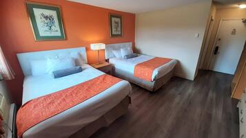 A hotel room with two beds, each with a white headboard, a red-orange bedspread, and a matching pillow. There are framed pictures on the orange wall, a bedside lamp, and a door with a lock.