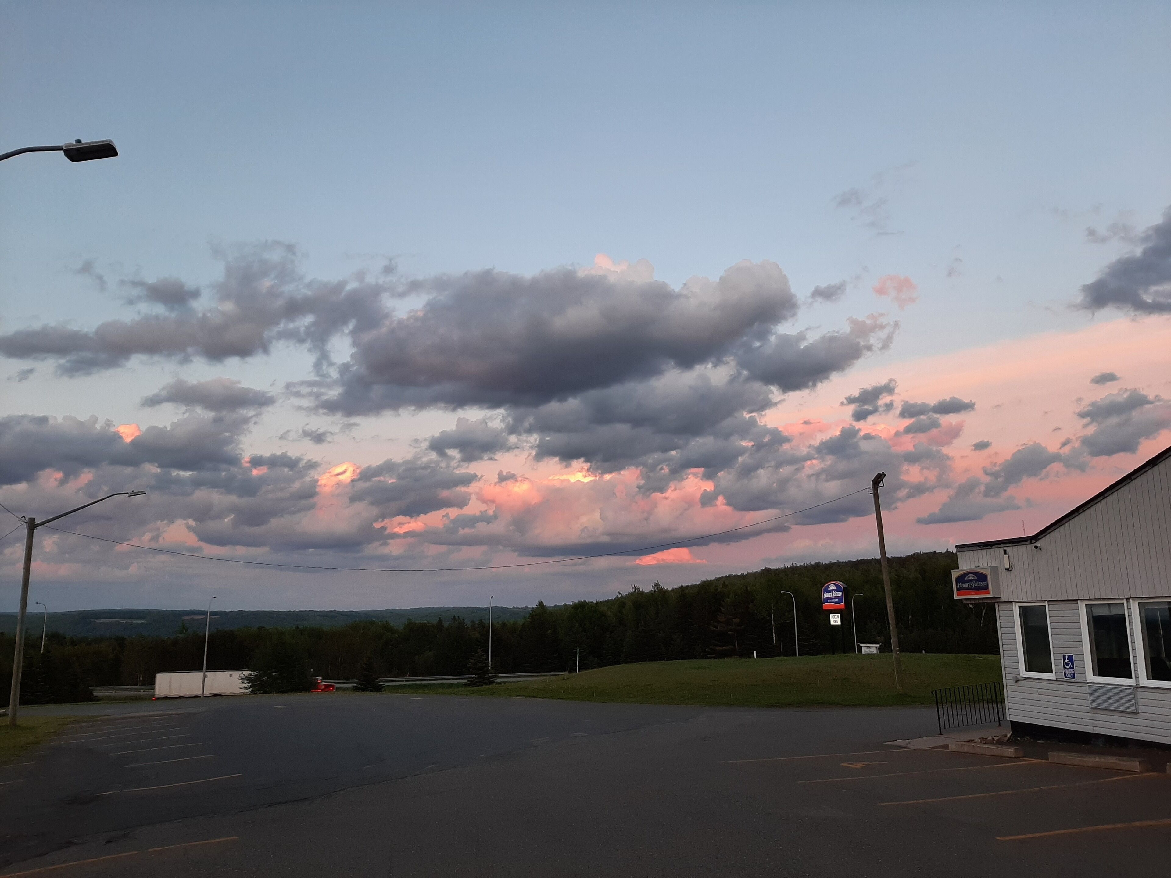A parking lot with a building on the right, a clear sky with clouds, and a distant view of trees and hills.