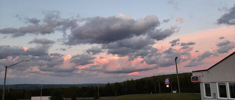 A parking lot with a building on the right, a clear sky with clouds, and a distant view of trees and hills.