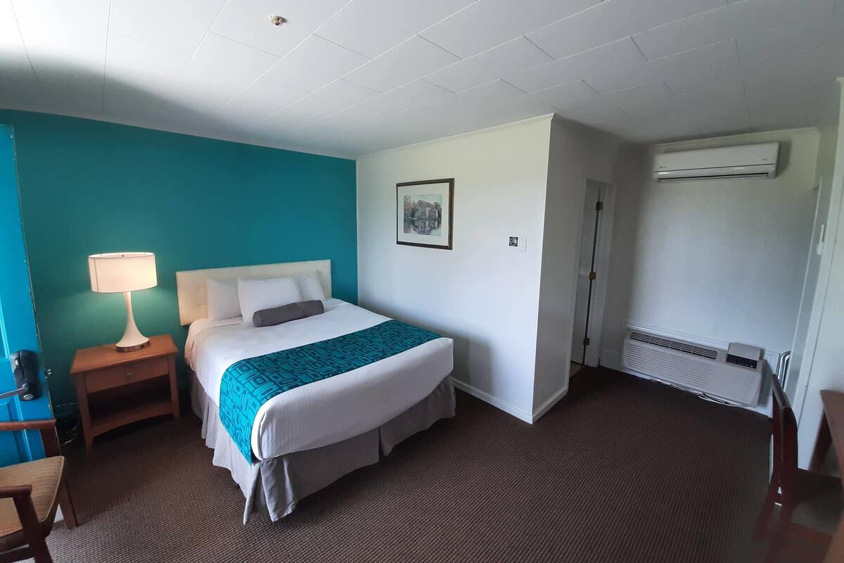 A hotel room with a bed, a nightstand, a lamp, a picture on the wall, and an air conditioning unit.