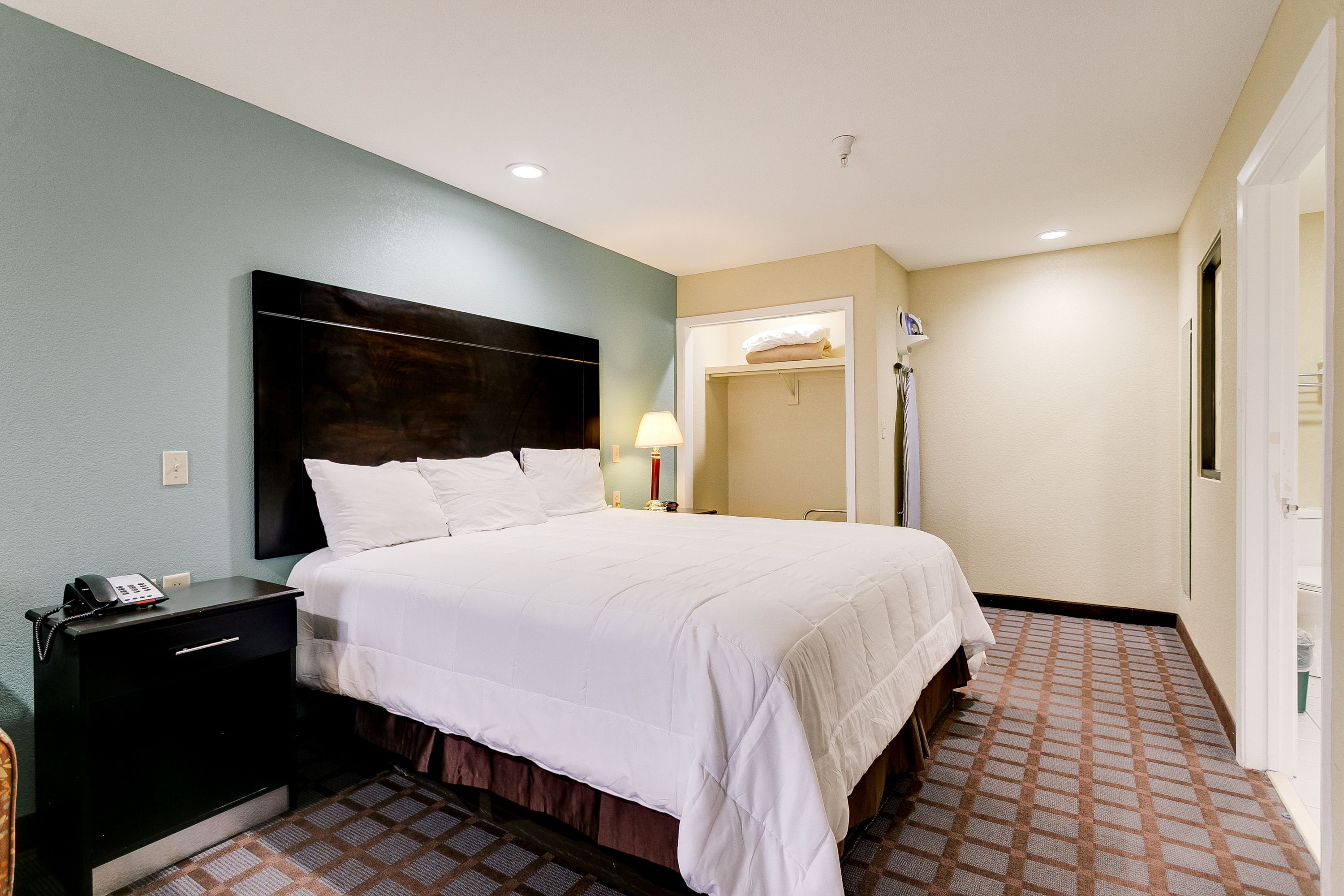 A hotel room with a large bed, a nightstand with a phone, a wall-mounted headboard, a closet with a towel, and a patterned floor.
