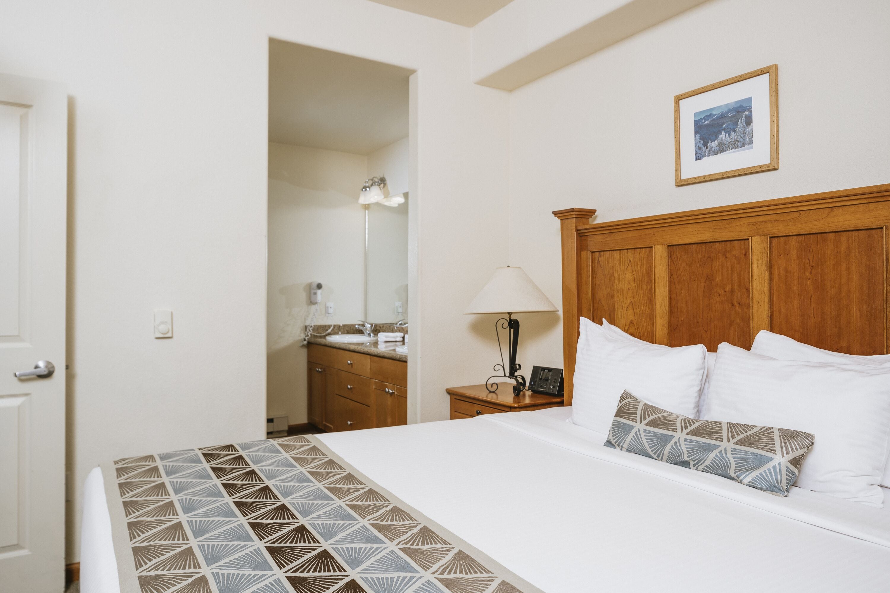 A hotel room with a bed, a wooden headboard, a nightstand, a lamp, a framed picture on the wall, and a bathroom visible through an open door.