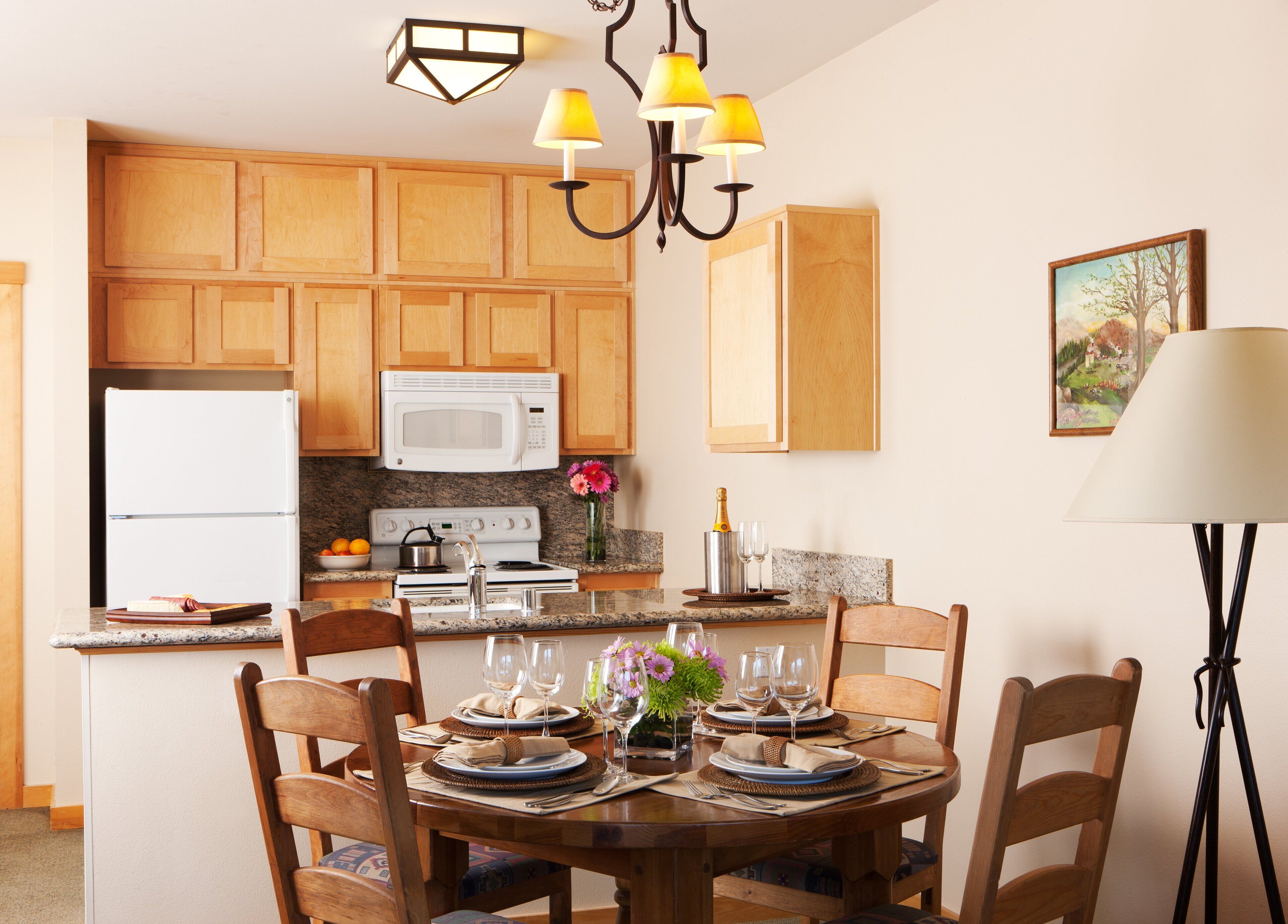 A kitchen with wooden cabinets, a white refrigerator, a microwave, and a stove. There is a dining table set with plates, glasses, and a vase of flowers.