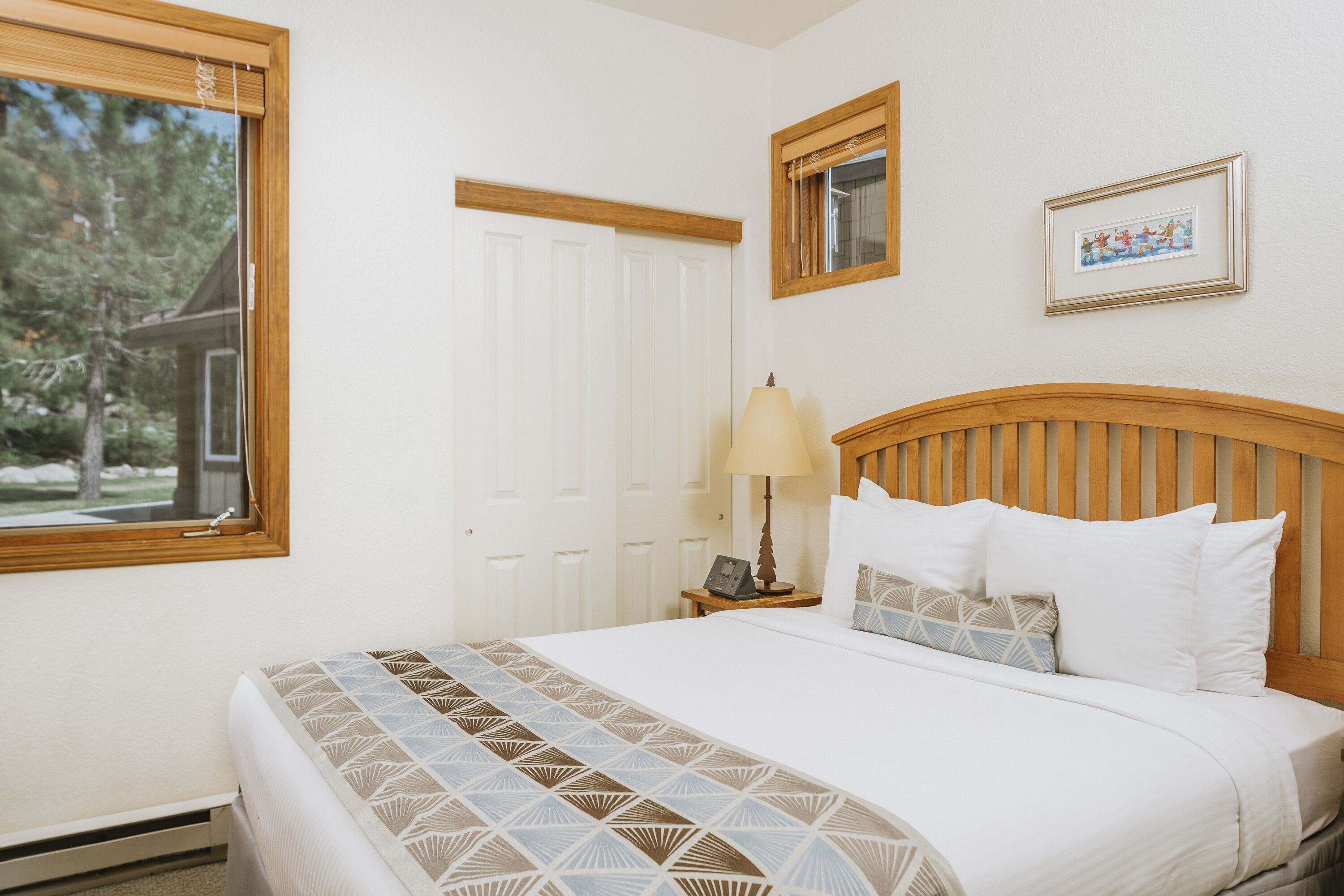 A bedroom with a large bed, a nightstand with a lamp, a framed picture on the wall, and a window with a view of trees.