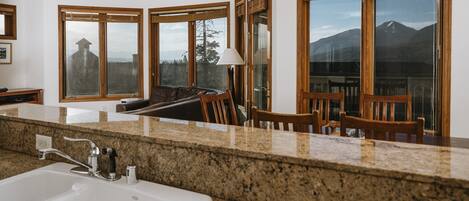 A kitchen with granite countertops, a white sink, wooden chairs, and large windows with a mountain view.