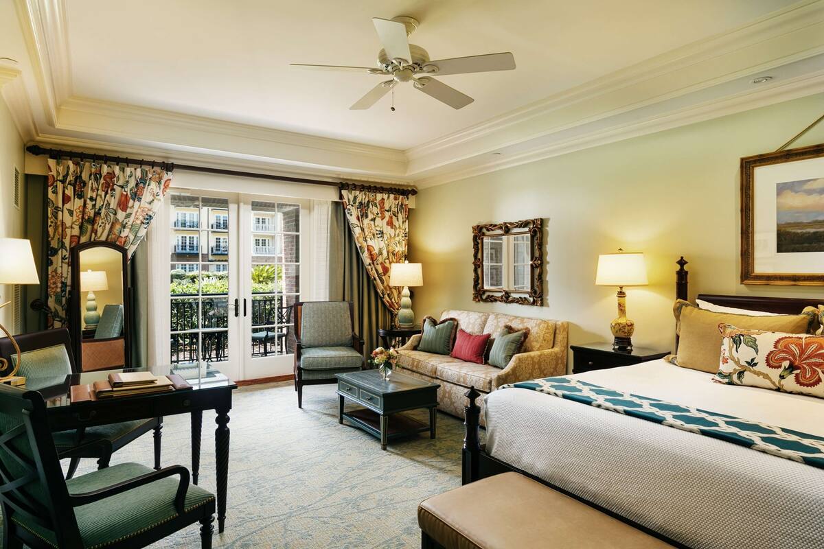 A hotel room with a bed, a sofa, a chair, a coffee table, and a desk. There is a balcony with a view of buildings.
