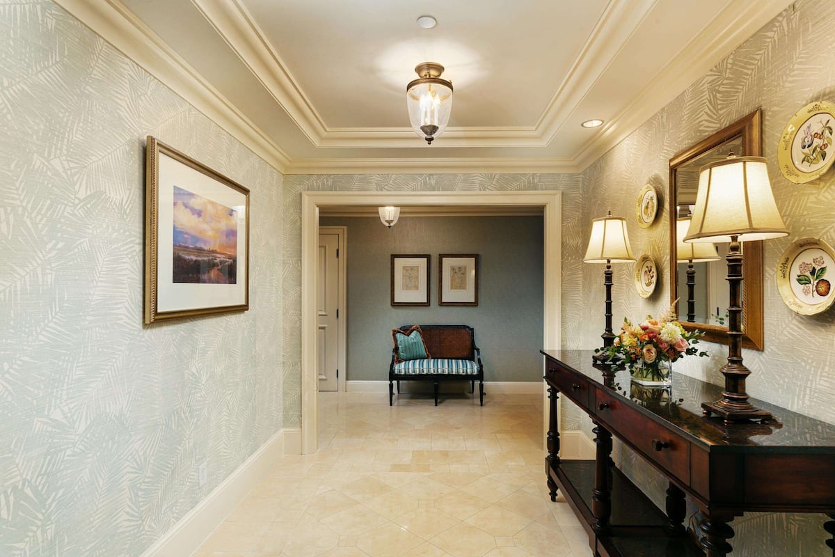 A hallway with patterned wallpaper, a bench, framed pictures, and a console table with lamps and decorative plates.