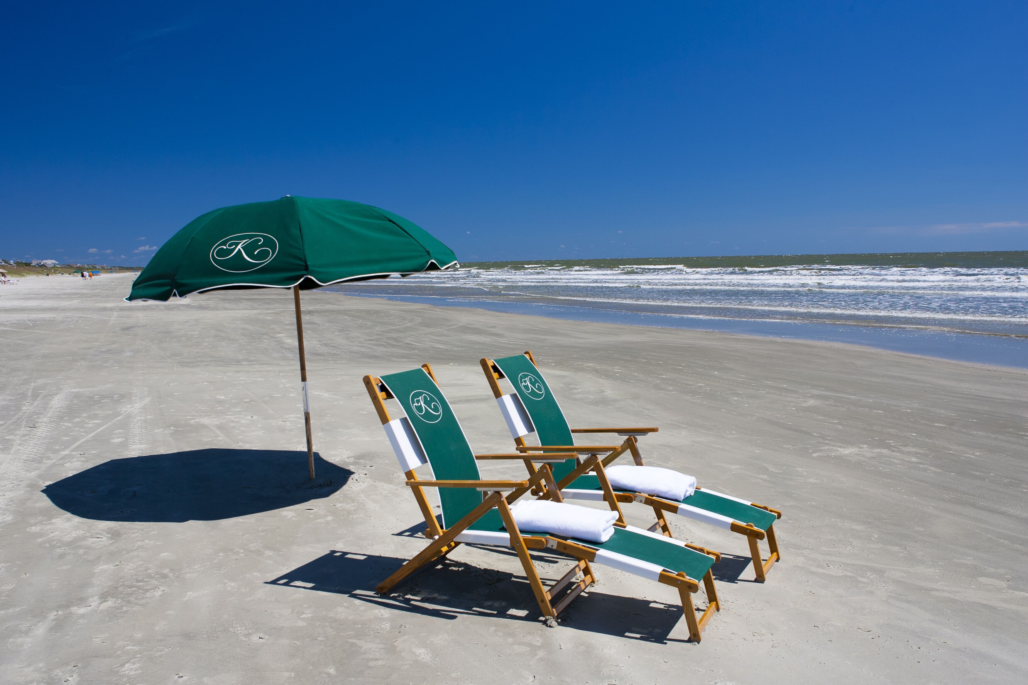 Two beach chairs with green towels and a matching umbrella on a sandy beach.