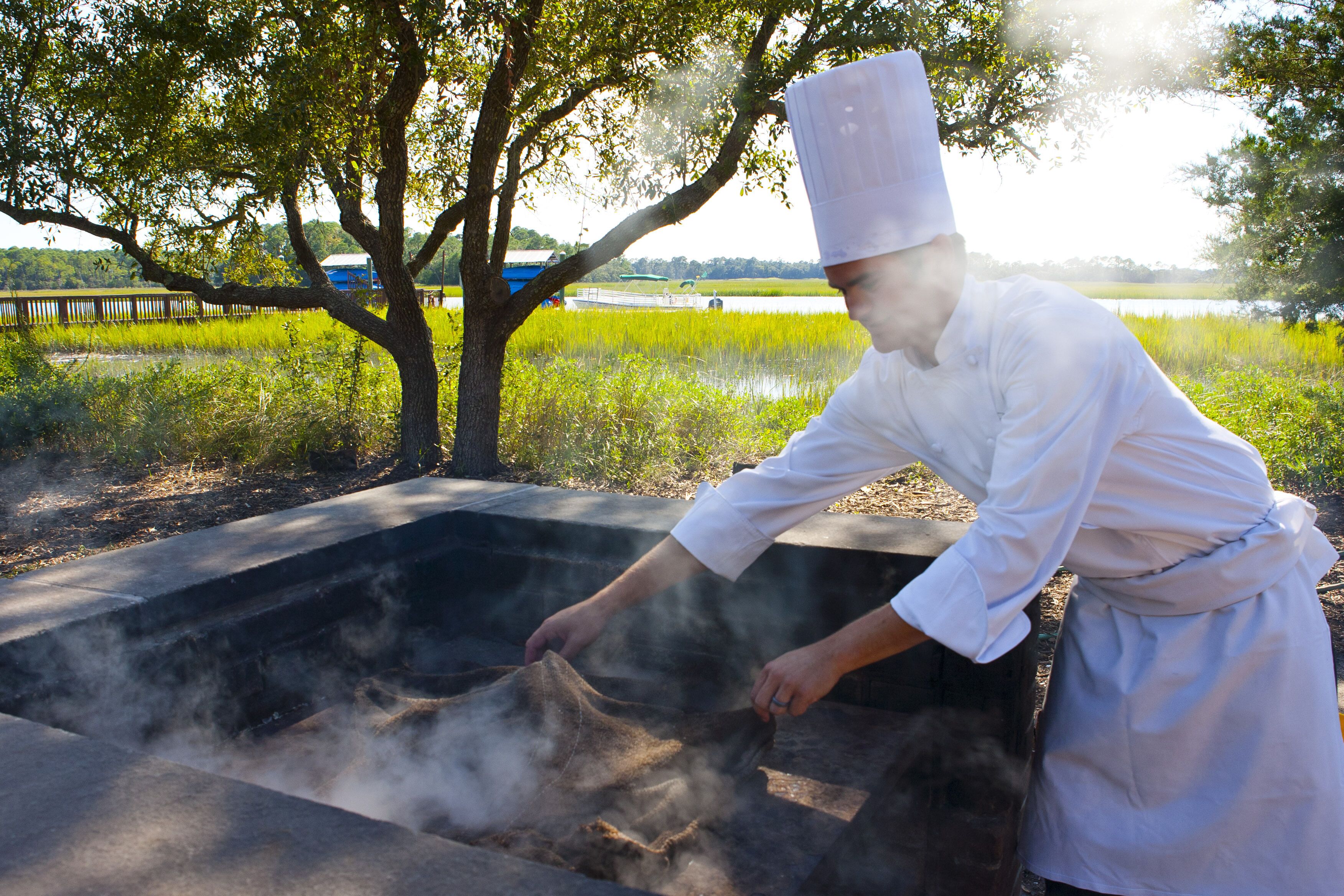 A chef cooking outdoors with a large pot on a stove, surrounded by trees and a body of water in the background.
