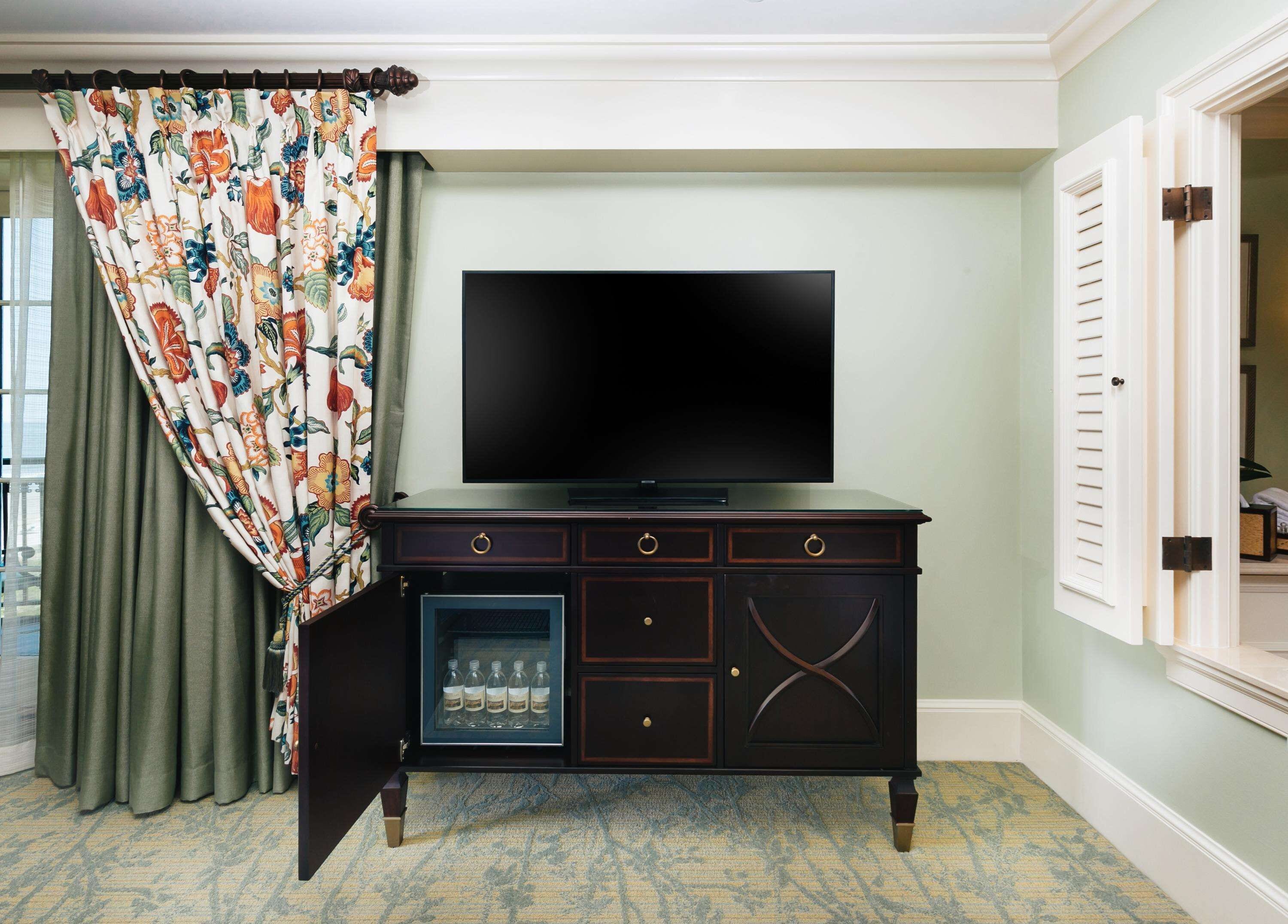 A room with a dark wooden TV stand, a flat-screen TV, a glass-fronted cabinet with bottles, a patterned curtain, and a green carpet.