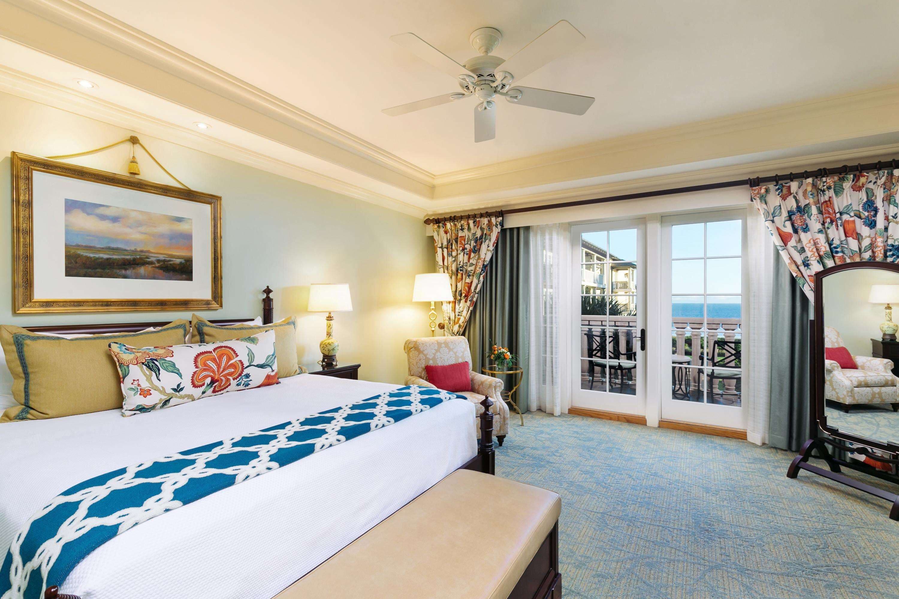 Une chambre d’hôtel avec un grand lit, un ventilateur de plafond, un balcon avec vue et un tableau au mur.