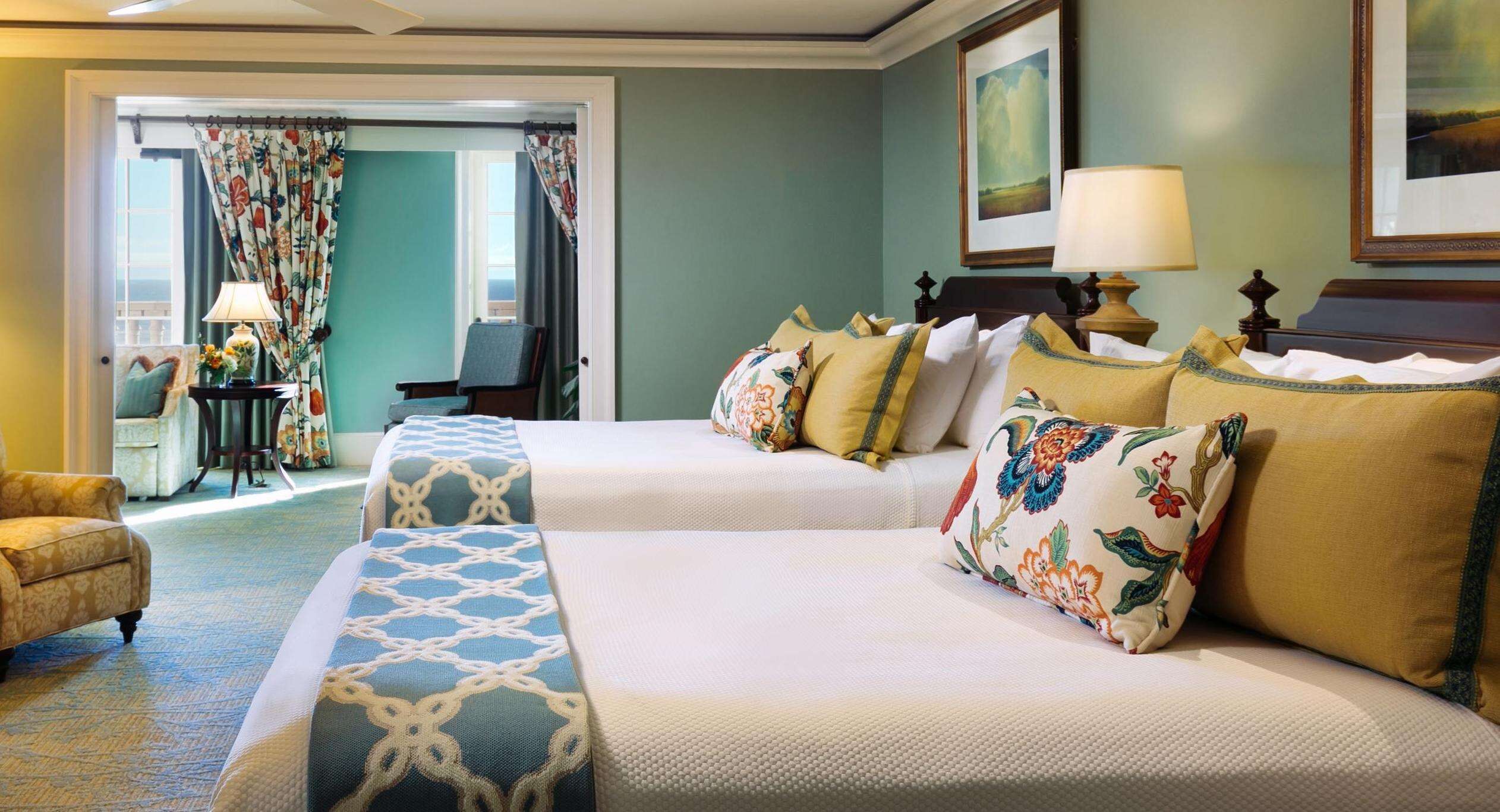 Quarto de hotel com duas camas, área de estar com uma cadeira, mesa e vista para o exterior.