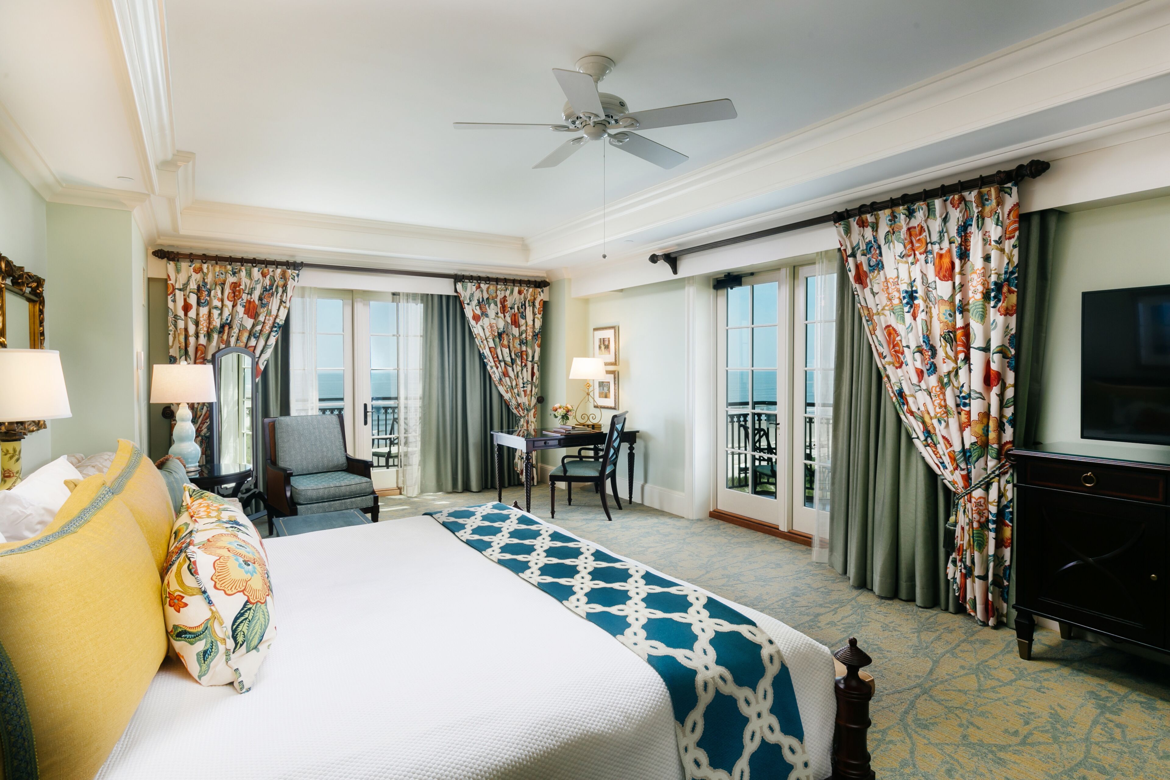 A hotel room with a large bed, a desk, a chair, a television, and a balcony with curtains.