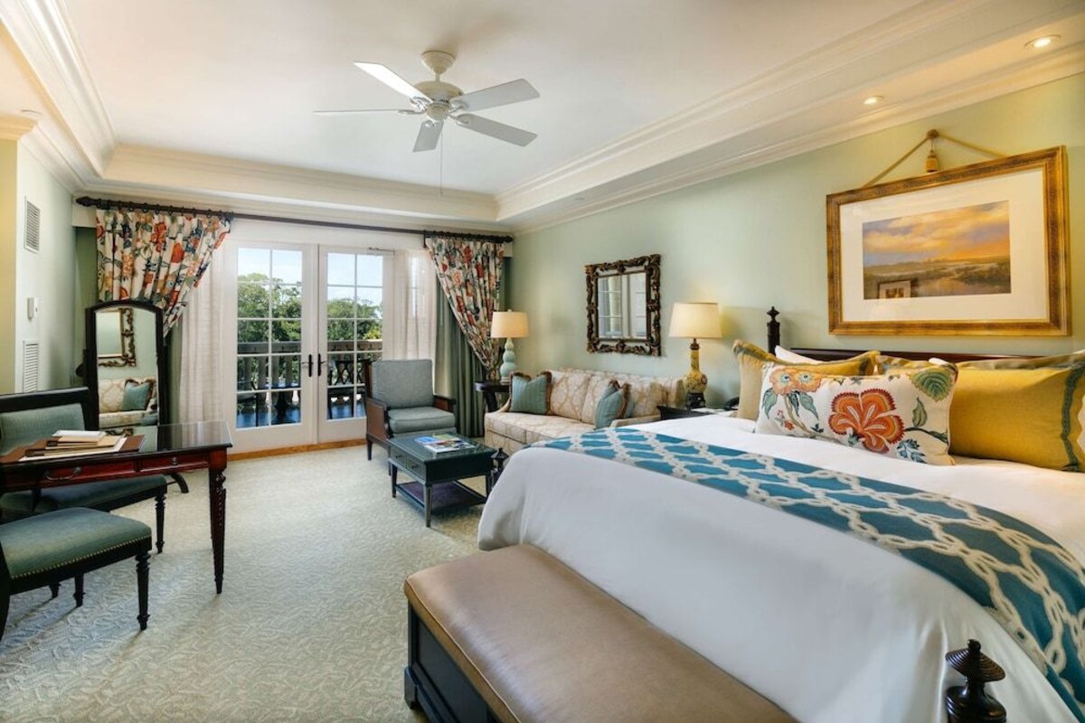Une chambre d’hôtel avec un lit, un canapé, un bureau, une chaise, un ventilateur de plafond et une grande fenêtre.