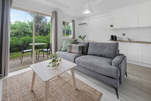 Una sala moderna con un sofá gris, una mesita con una planta y un ventanal corredero que da a un patio con mobiliario exterior.