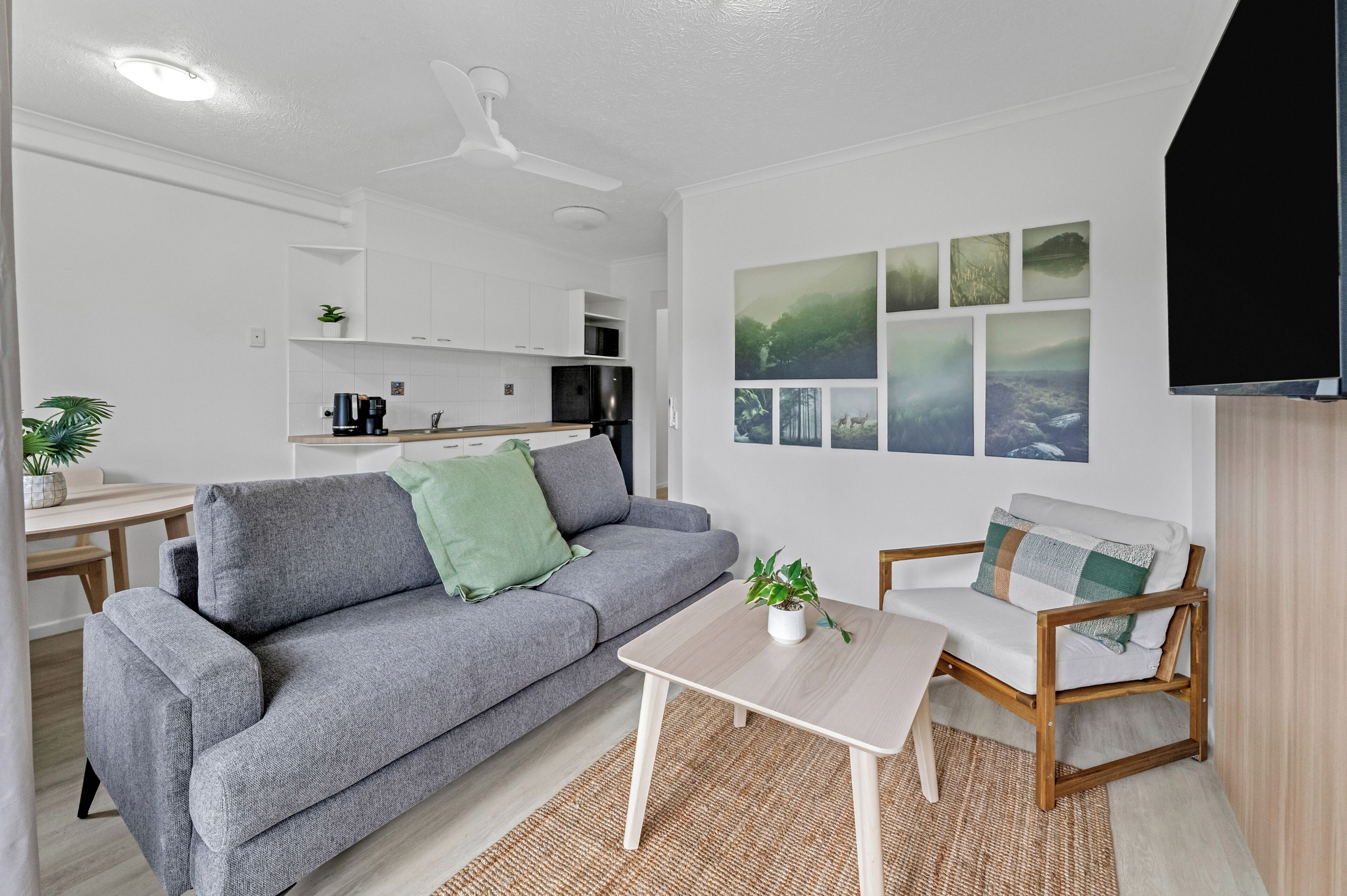 A modern living room with a grey sofa, a wooden coffee table, and a chair with a cushion. A flat-screen TV is mounted on the wall.