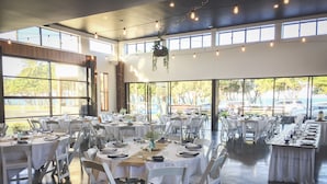 A spacious indoor venue with multiple round tables set for dining, large windows offering views of the outdoors, and hanging lights. - Mackay Seabreeze Apartments (Slade Point)