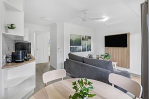 Una sala de estar moderna con un sofá gris, una mesa de comedor con sillas, una zona de cocina con cafetera y un televisor de pantalla plana montado en la pared.
