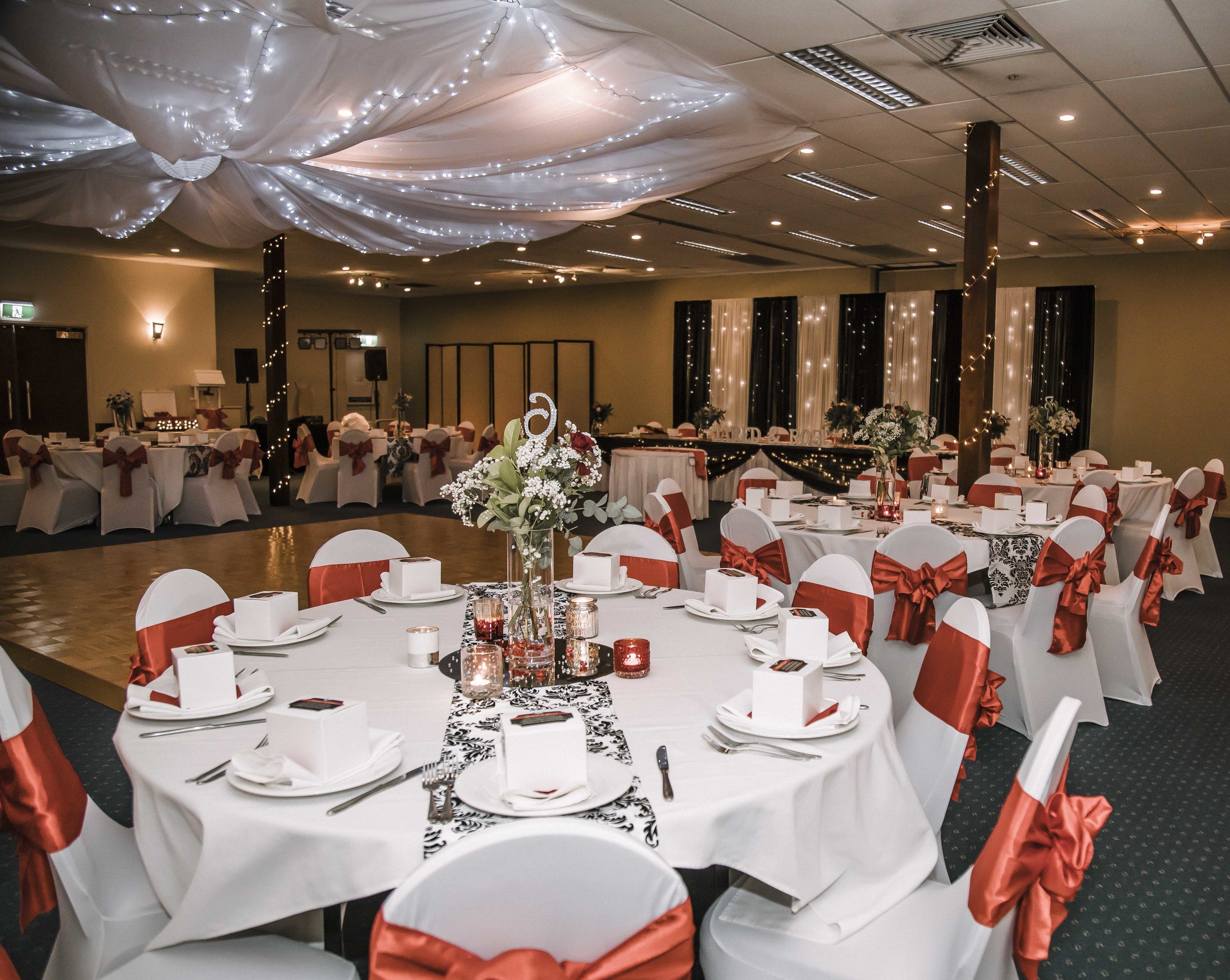 A banquet hall with round tables set for a formal event, featuring white tablecloths, red sashes, and floral centerpieces.