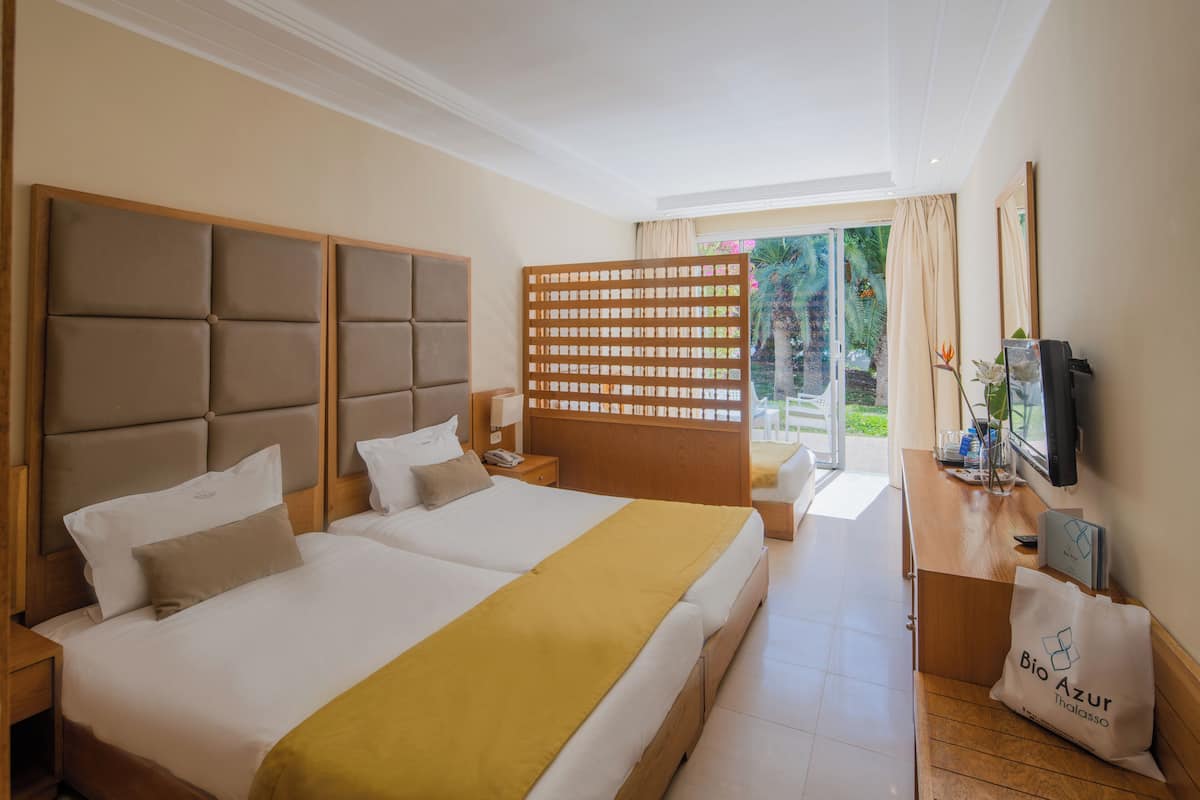 Habitación de hotel con dos camas, cabecero de madera, escritorio con computadora, televisión y puerta corrediza de vidrio al exterior.