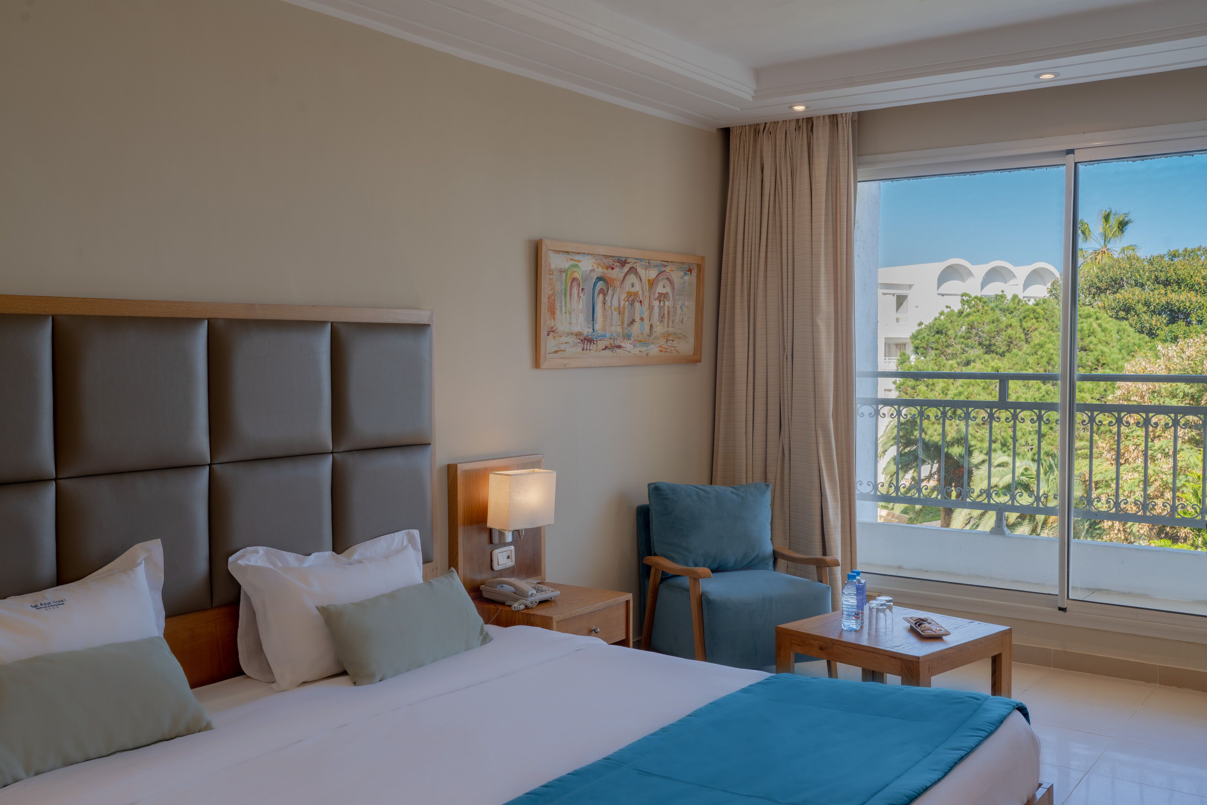 Une chambre d’hôtel avec un grand lit, un bureau, une chaise, une table de chevet et une fenêtre donnant sur un paysage verdoyant et des bâtiments.