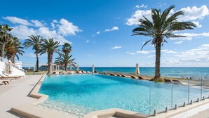 Piscina azul cristalina com palmeiras e espreguiçadeiras, rodeada por um deck de azulejos e vista para o mar.