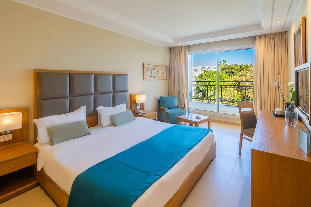 Un cuarto de hotel con una cama grande, un escritorio, una silla, un balcón con vistas y un cuadro en la pared.