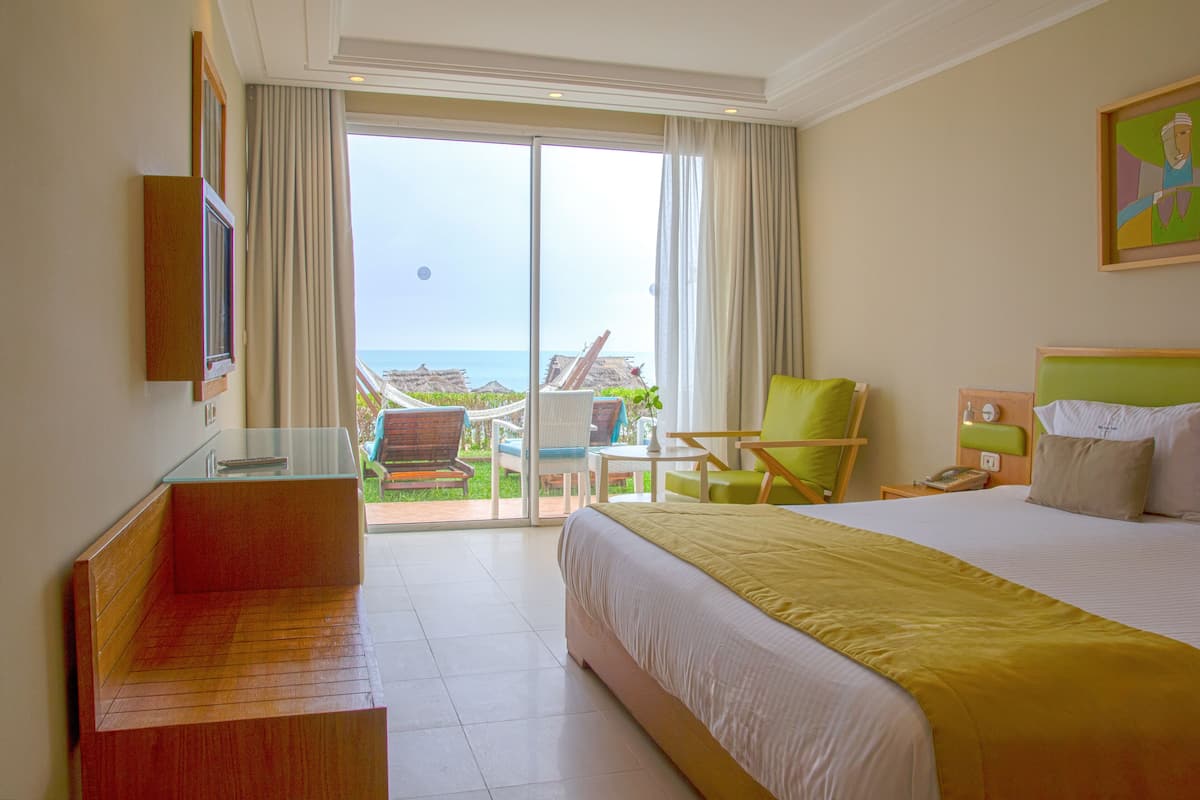 Habitación de hotel con una cama grande, un banco de madera, una silla, una mesita y vista al océano a través de un ventanal.