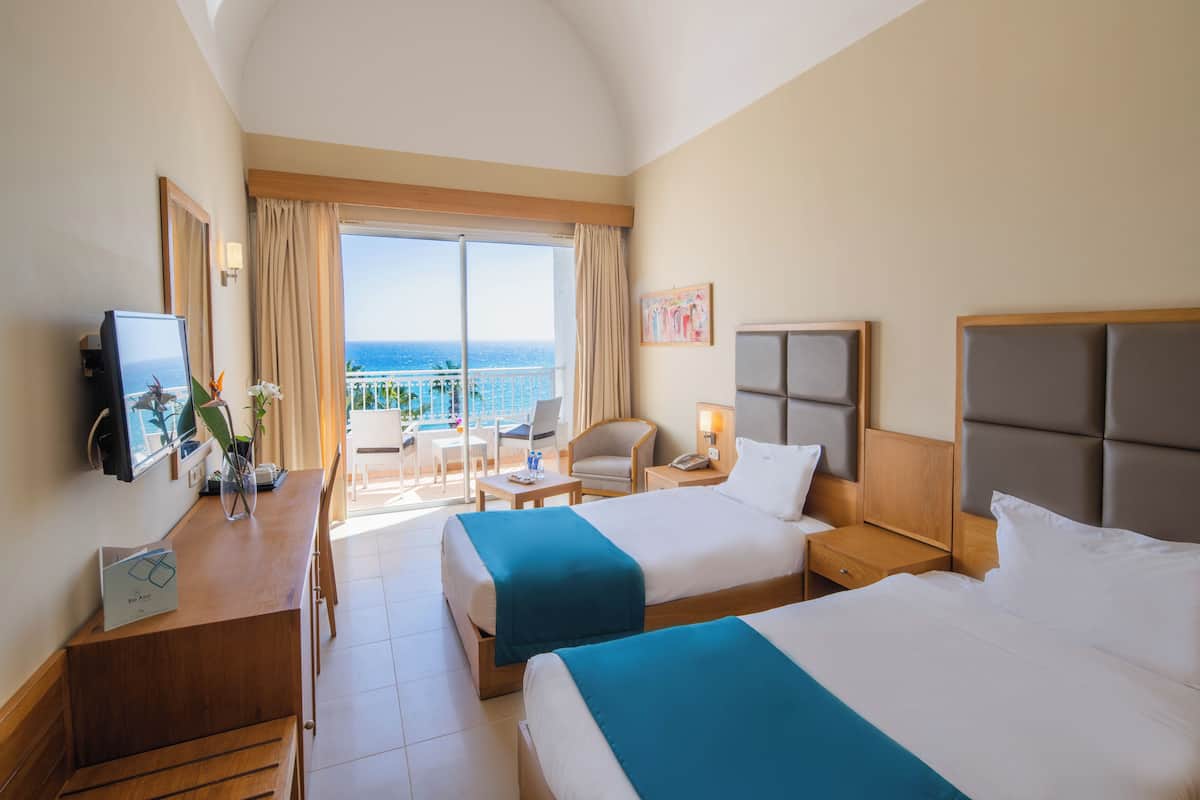 Habitación de hotel con dos camas, un escritorio, una silla, televisión y balcón con vista al mar.