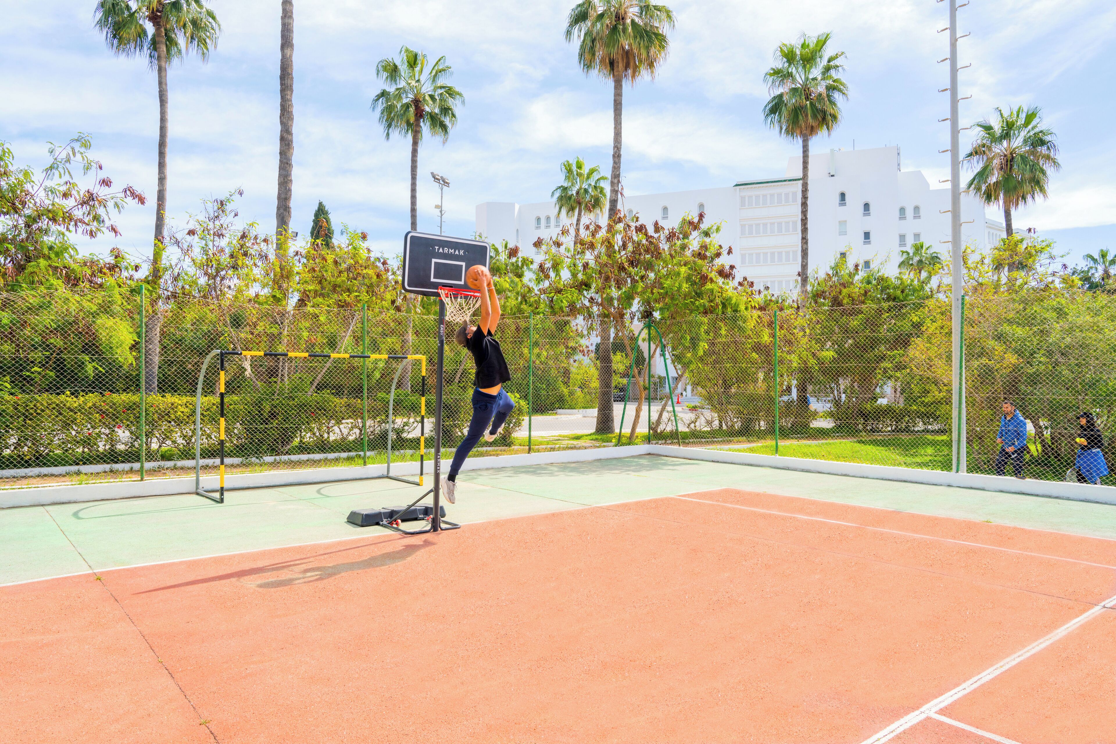 An outdoor basketball court with a net, palm trees, and a fence.