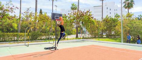 An outdoor basketball court with a net, palm trees, and a fence.