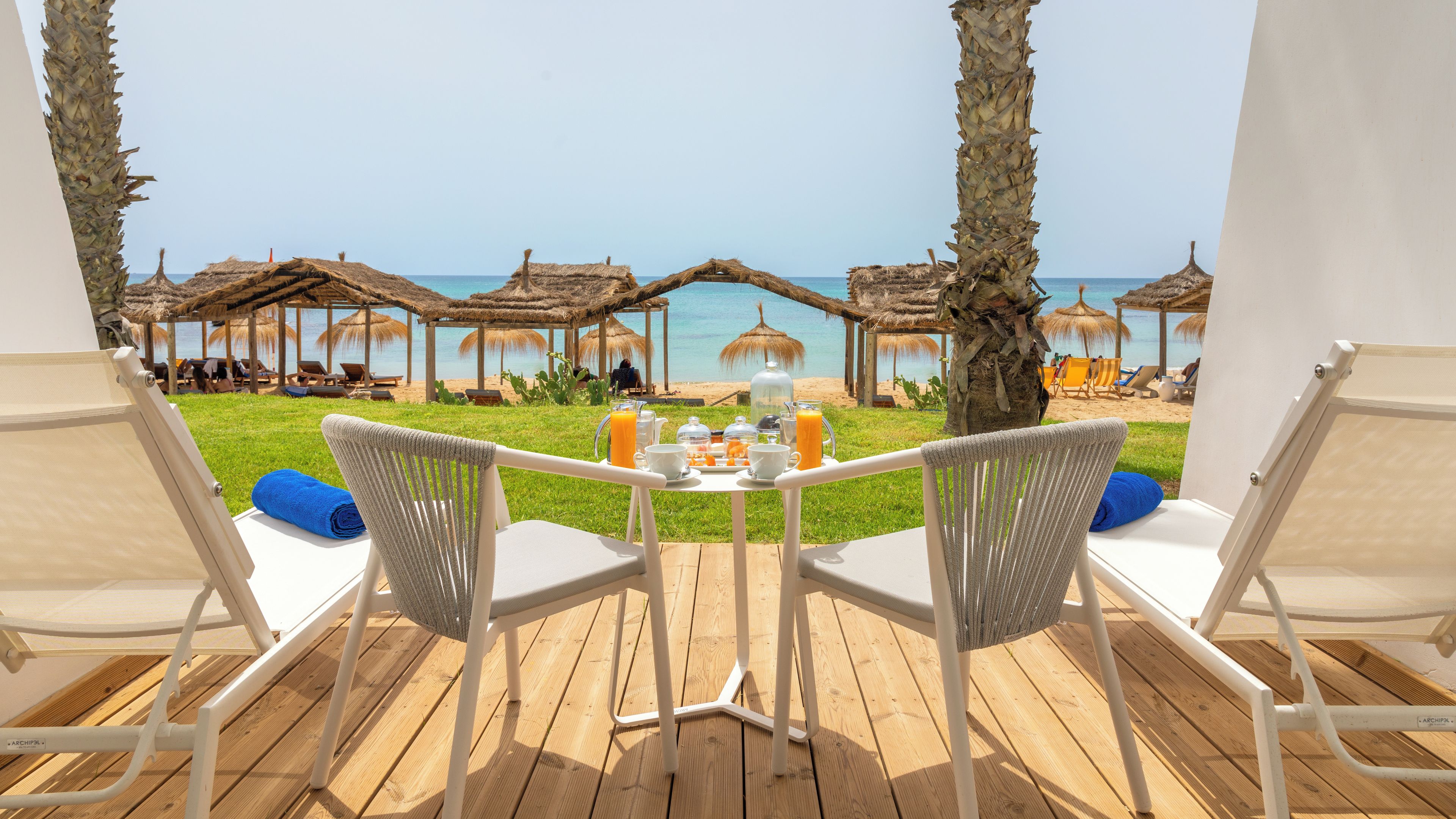 Terraza con dos sillas y una mesa dispuesta para el desayuno, con vista a una playa con sombrillas y sillas de descanso.