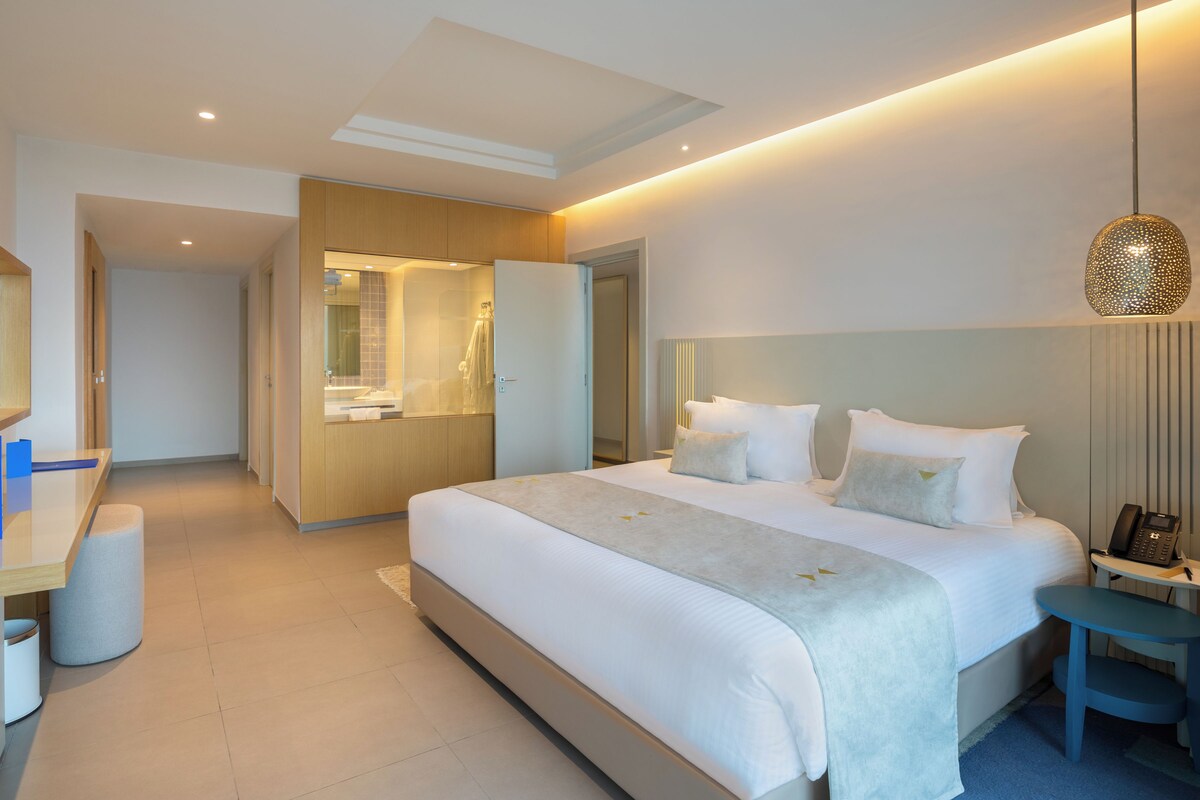 A modern hotel room with a large bed, a desk, a mirror, and a bathroom visible through an open door.