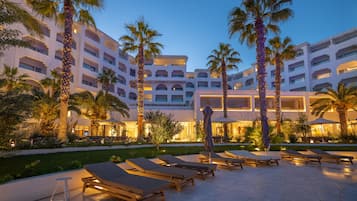 Une cour d’hôtel avec des chaises longues, des palmiers et une façade d’immeuble bien éclairée.