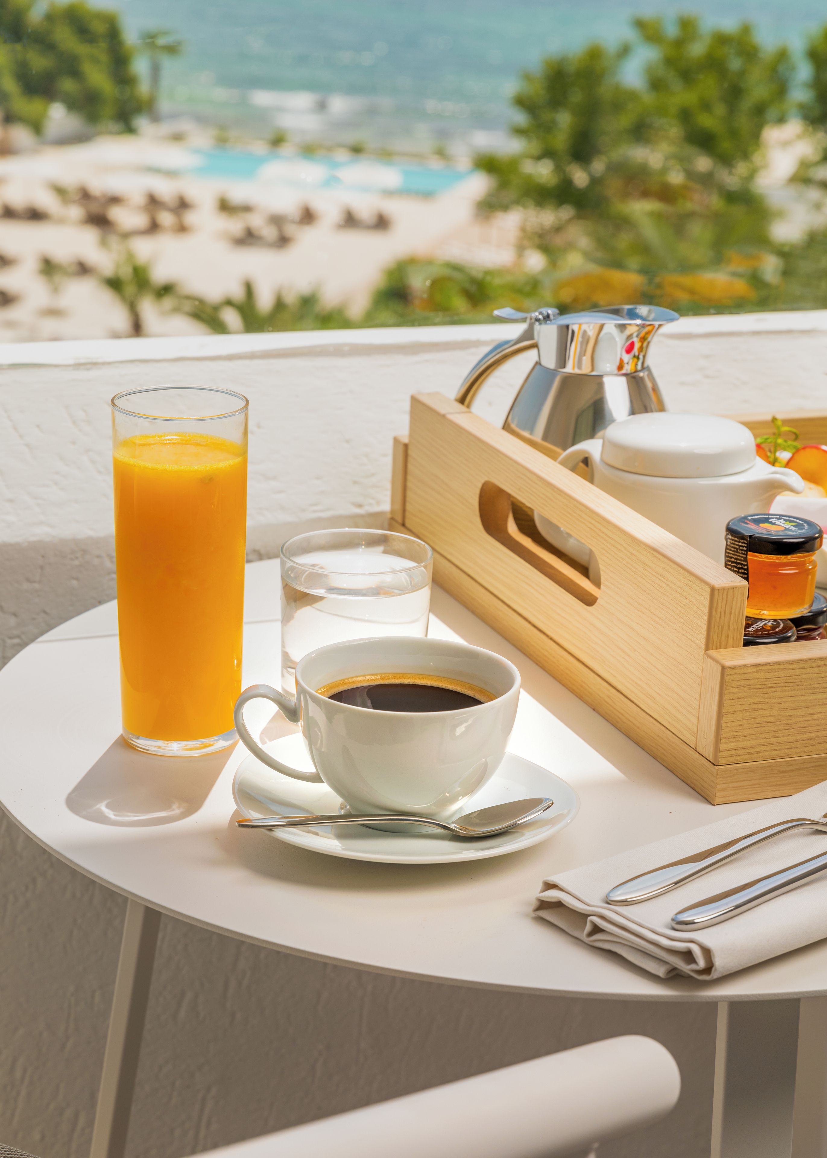 Una mesa con un vaso de jugo de naranja, una taza de café, un vaso de agua, un juego de té y una servilleta doblada en un balcón con vistas a una playa.