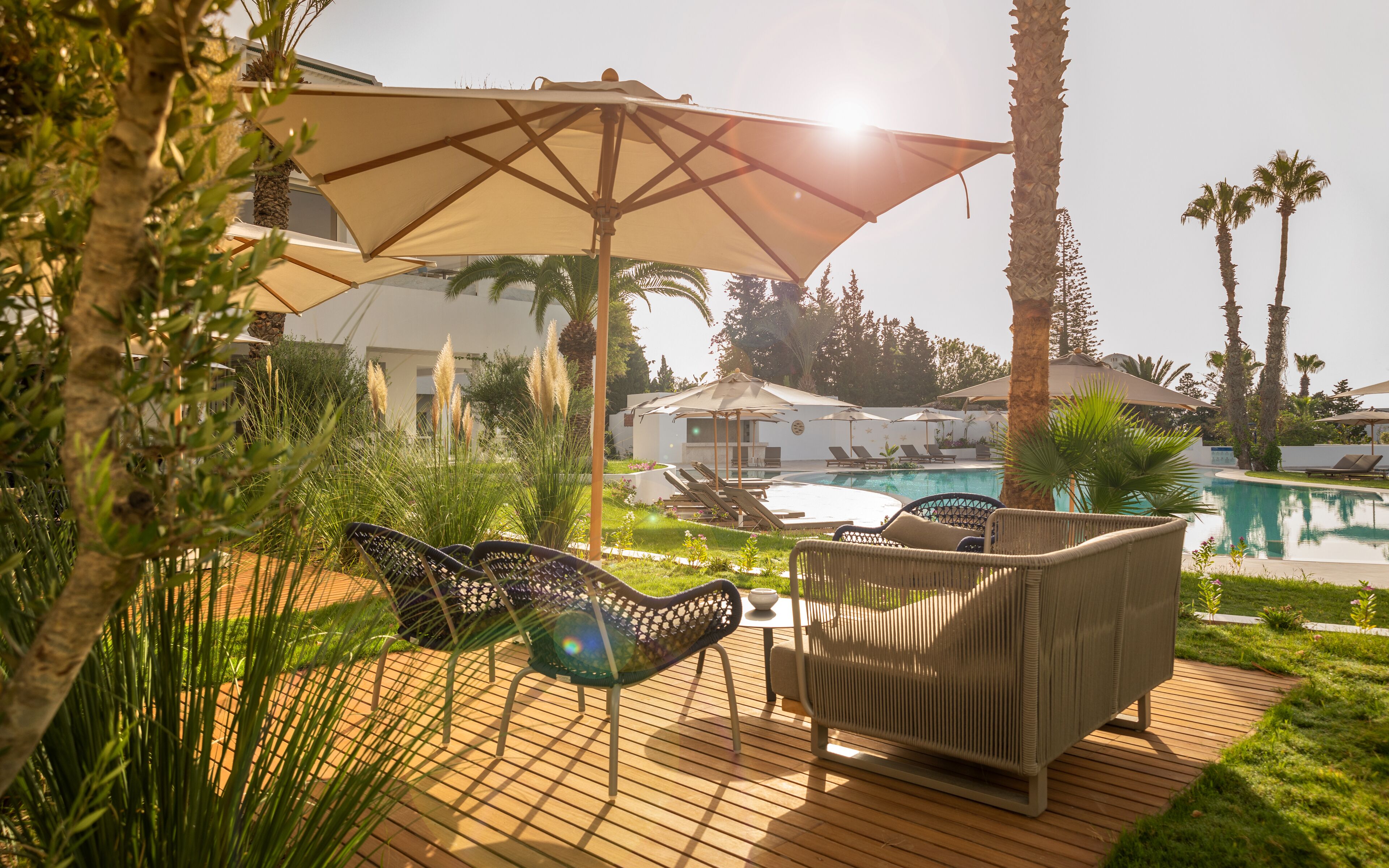 Zona junto a la piscina con sillones de descanso y sombrillas, rodeada de follaje y palmeras.