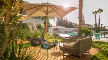 Zona junto a la piscina con sillones de descanso y sombrillas, rodeada de follaje y palmeras.