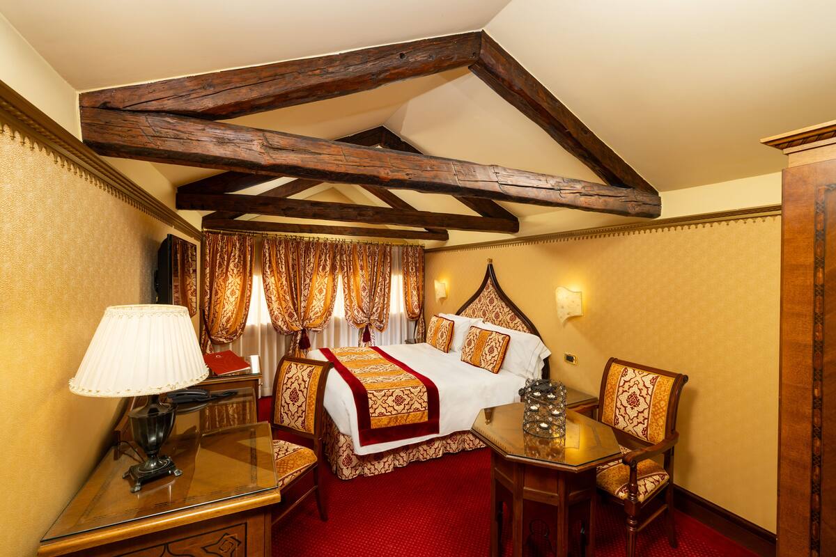 A hotel room with a wooden ceiling, a bed with a red and gold bedspread, a table with a lamp, and a chair.