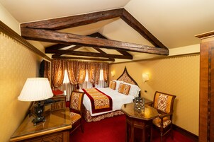 A hotel room with a wooden ceiling, a bed with a red and gold bedspread, a table with a lamp, and a chair.