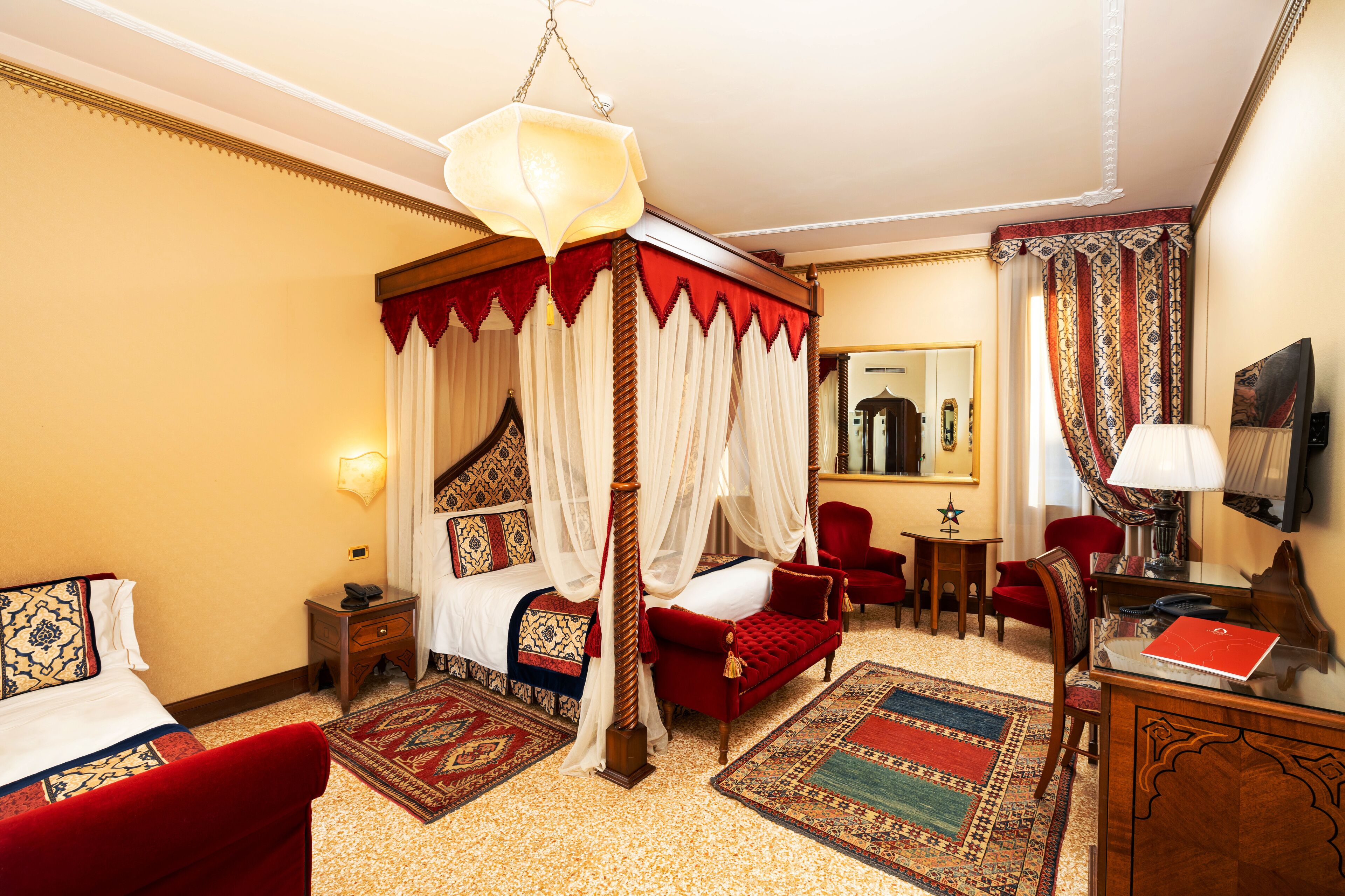 Uma cama com dossel e cortina, um sofá vermelho, uma escrivaninha de madeira com abajur e uma poltrona vermelha em um quarto de hotel.