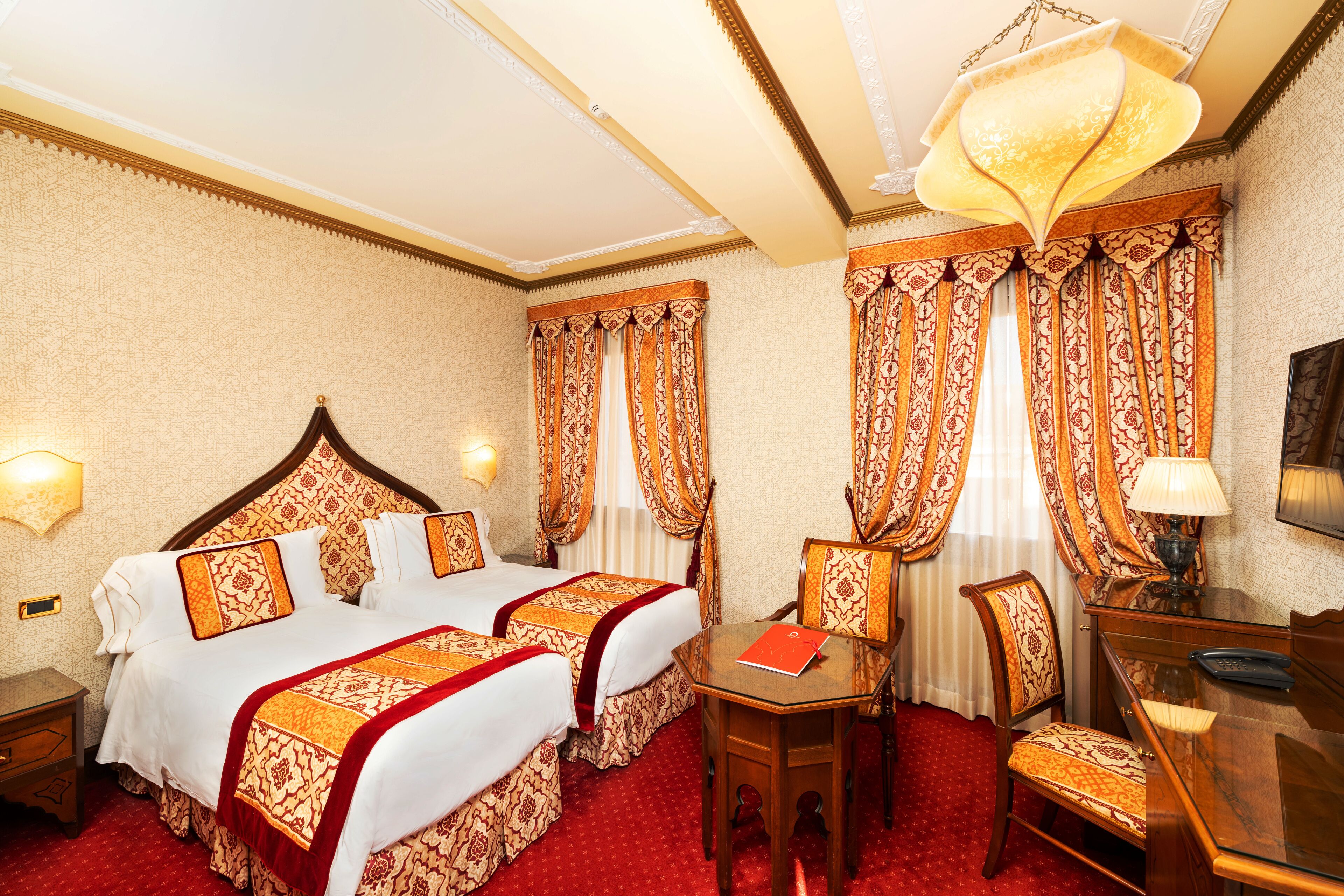 A hotel room with two beds, a desk, a chair, and a lamp.
