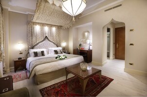 A spacious bedroom with a large bed, a wooden nightstand, a desk, and a mirror. There is a red patterned rug on the floor.