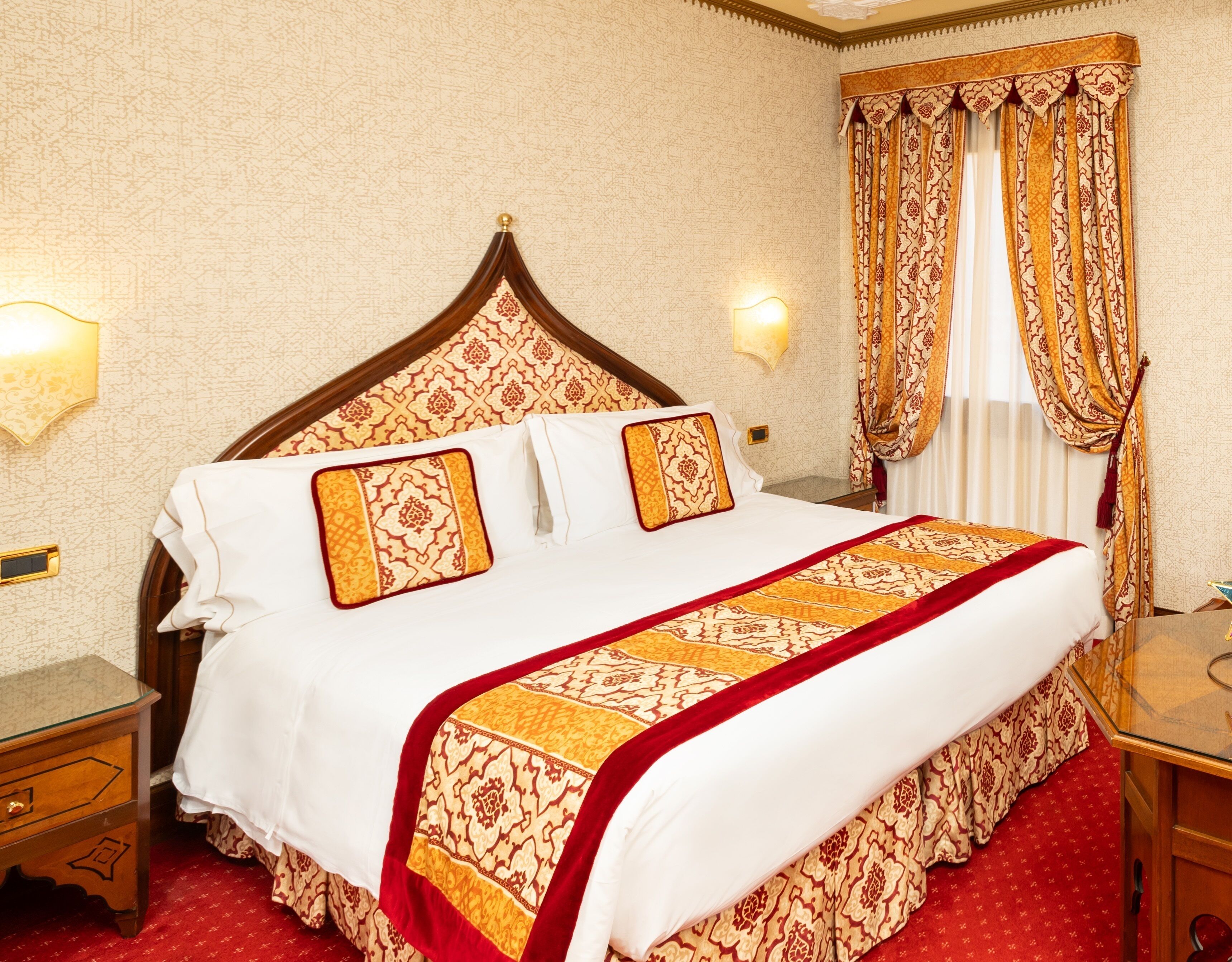 Ein Hotelzimmer mit einem großen Bett, einem Holz-Kopfteil, einem roten Teppich und einem gemusterten Vorhang.